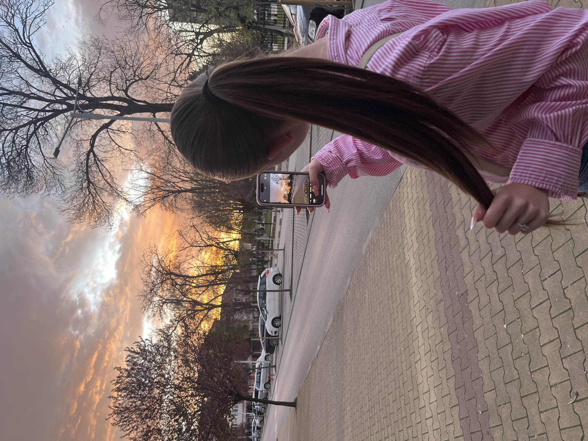A woman with long brown hair is taking a photo of a sunset with her smartphone. She is wearing a pink and white striped shirt. She is standing on a sidewalk paved with tan and brown bricks. A street runs parallel to the sidewalk with parked white cars. Trees line the street, silhouetted against the sunset sky. The sky is a mix of orange, yellow, and gray clouds. The phone in her hand displays a similar image of the sunset. The location is most likely in the city of Nova Zagora, Bulgaria.