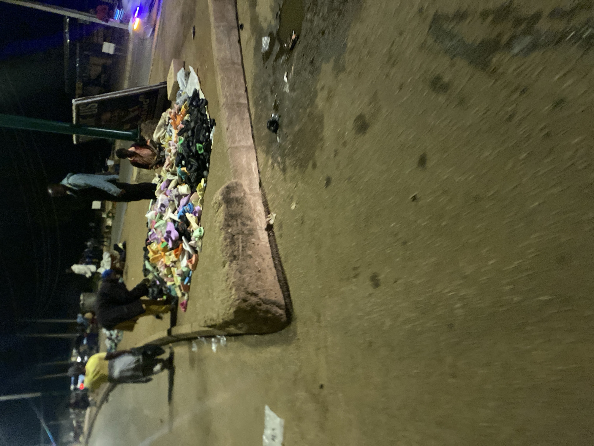 In Bukuru, Nigeria, a dimly lit street scene unfolds at night. A pile of assorted shoes, some colorful and others black, is scattered on the sidewalk. Two individuals are visible near the shoes: one wearing a dark shirt and pants bends over the pile, while another person stands nearby. A third individual in a light-colored shirt and dark pants stands further back, and a child in a pink shirt sits on the ground beside a sign with text that reads "US HAND." In the background, other figures and a vehicle are blurred, suggesting activity on the street. The ground surface appears wet and dirty, with scattered debris.