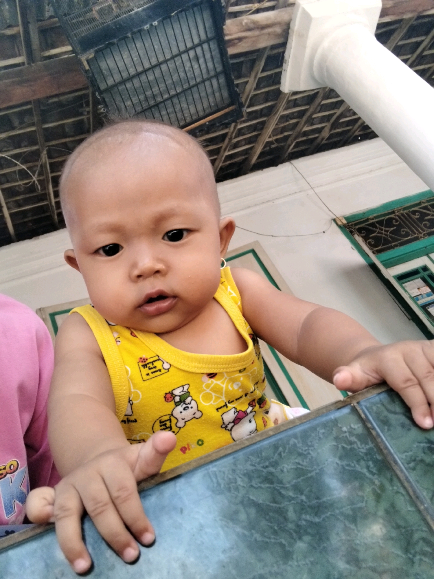 The image shows a close-up, low-angle, medium shot of a bald infant wearing a yellow sleeveless shirt with a cartoon bear print. The infant is positioned in the foreground, leaning forward, with their hands resting on a dark green, patterned surface. The infant's face is turned towards the camera, with dark eyes and slightly parted lips. To the left of the infant, a portion of a pink garment is visible.
In the background, there is a white, fluted pillar and the underside of a thatched roof structure with exposed wooden beams and a dark metal cage-like object. The environment appears to be outdoors, possibly a porch or veranda, with hints of a building structure with green trim and a window with wrought iron detailing. The overall setting suggests a residential area in Kepuh Kiriman, Indonesia. The lighting indicates daytime.