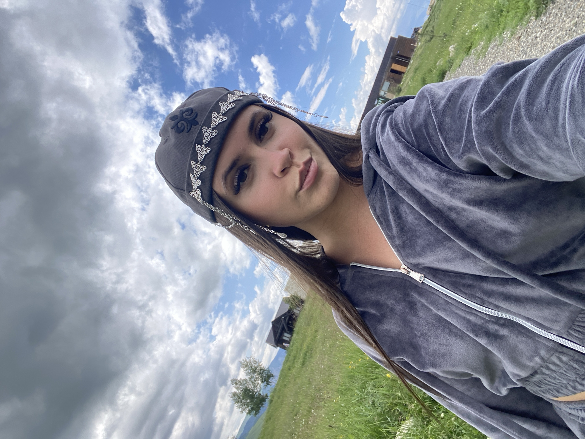 A young woman is taking a selfie outdoors in a grassy, hilly area. She is wearing a grey velour tracksuit and a grey beanie with decorative silver chain embellishments and an emblem. Her long, dark hair is falling forward. The background shows a cloudy sky, green hills with scattered trees, and a modern wooden building on a slope. A dirt path runs along the right side of the frame. The city of Tsalkita, Russia is not visible in this image, as the scene depicts a rural or mountainous landscape.