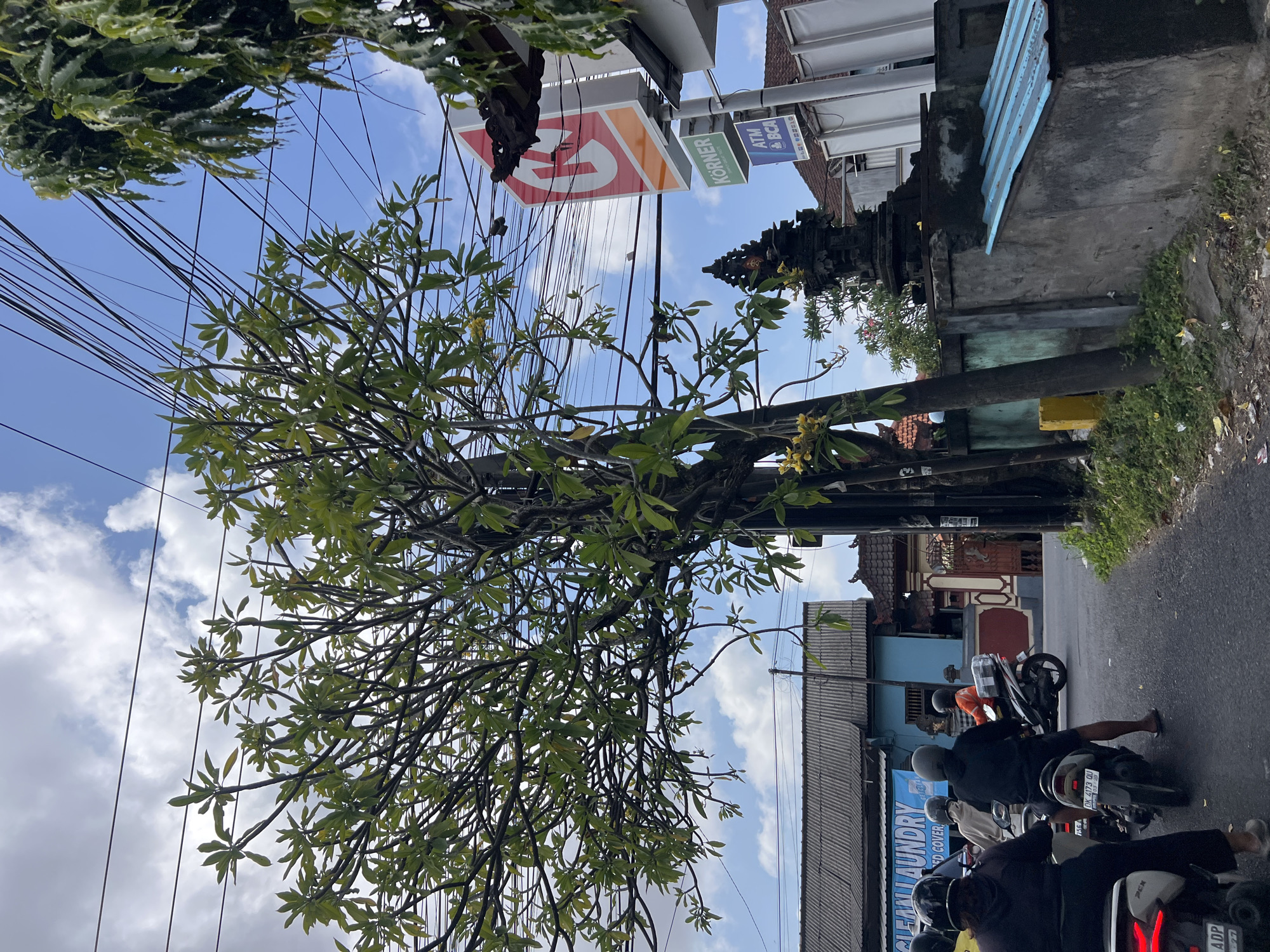 An eye-level, full shot captures a scene on a paved road in Denpasar, Indonesia, where several people are riding motorcycles. A large tree with green foliage and some yellow flowers dominates the foreground, its branches reaching across overhead power lines. In the background, a blue building with a corrugated metal roof is visible, alongside a red and white sign for Circle K. Further to the right, a smaller sign reads "KORNER" and below it, "ATM BCA." A Balinese shrine, a dark stone structure adorned with decorative carvings, sits on a grey concrete wall to the right of the road, partially obscured by the tree. The sky above is blue with scattered white clouds. The road surface appears worn, with some debris and grass growing along the sides. The overall impression is of a typical street scene in a tropical urban environment.