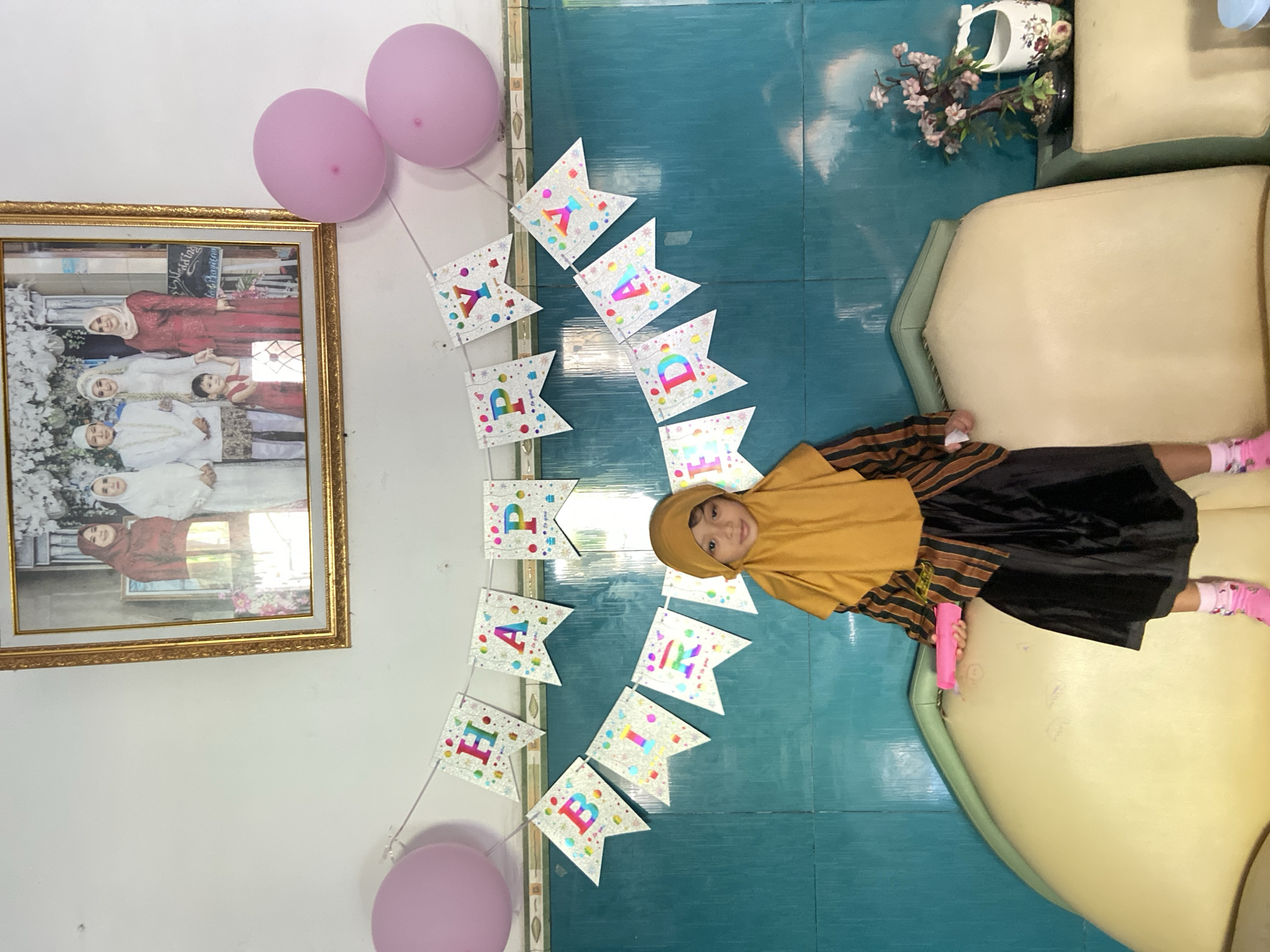 A small girl is laying on a light-colored sofa inside a home. The girl is wearing a light brown hijab, a striped shirt, and a black skirt, along with pink socks. She's holding a pink object. The background consists of blue tiled flooring, a white wall with a framed picture of a group of women, pink balloons, and a "Happy Birthday" banner attached to the wall. The location is Gelam, Indonesia.