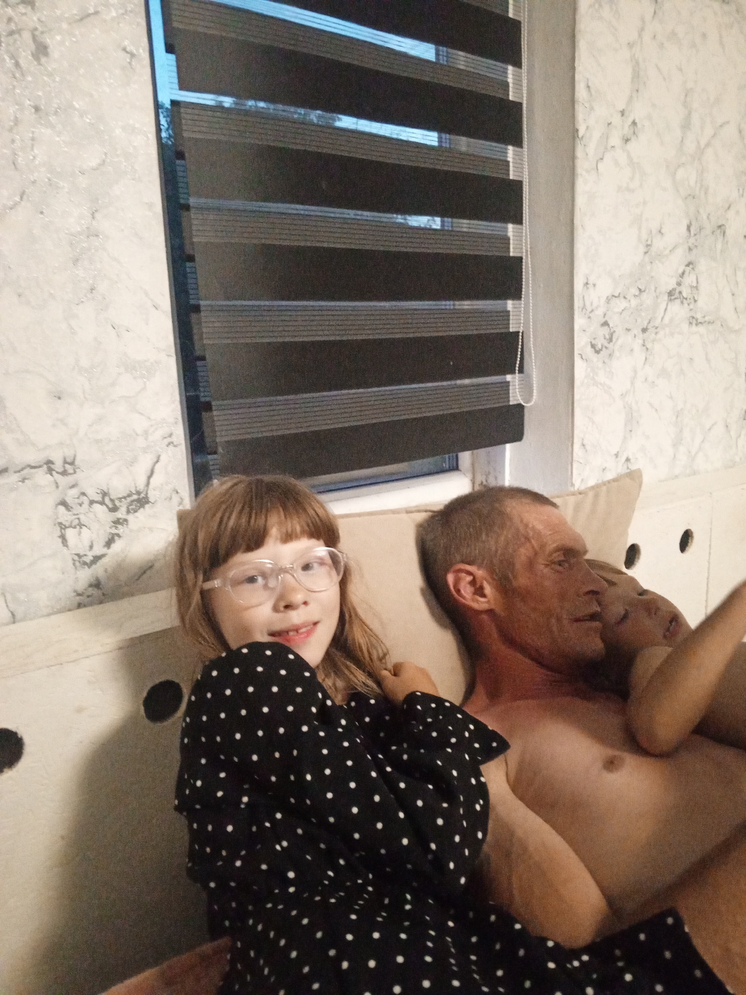 In a domestic indoor setting, likely a bedroom in Buraly, Russia, a family is gathered on a bed. A young girl with blonde hair and glasses, wearing a black and white polka dot dress, is in the foreground, facing the camera and smiling. She is positioned to the left of the frame. To her right, an adult male, appearing to be her father, is lying down, his body partially visible. He is looking to his right. Tucked close to the man, another child, likely younger, is also lying down, with their head resting on the man's chest. The background shows a marble-patterned wall and a window with horizontal blinds. The overall atmosphere suggests a casual, intimate family moment.