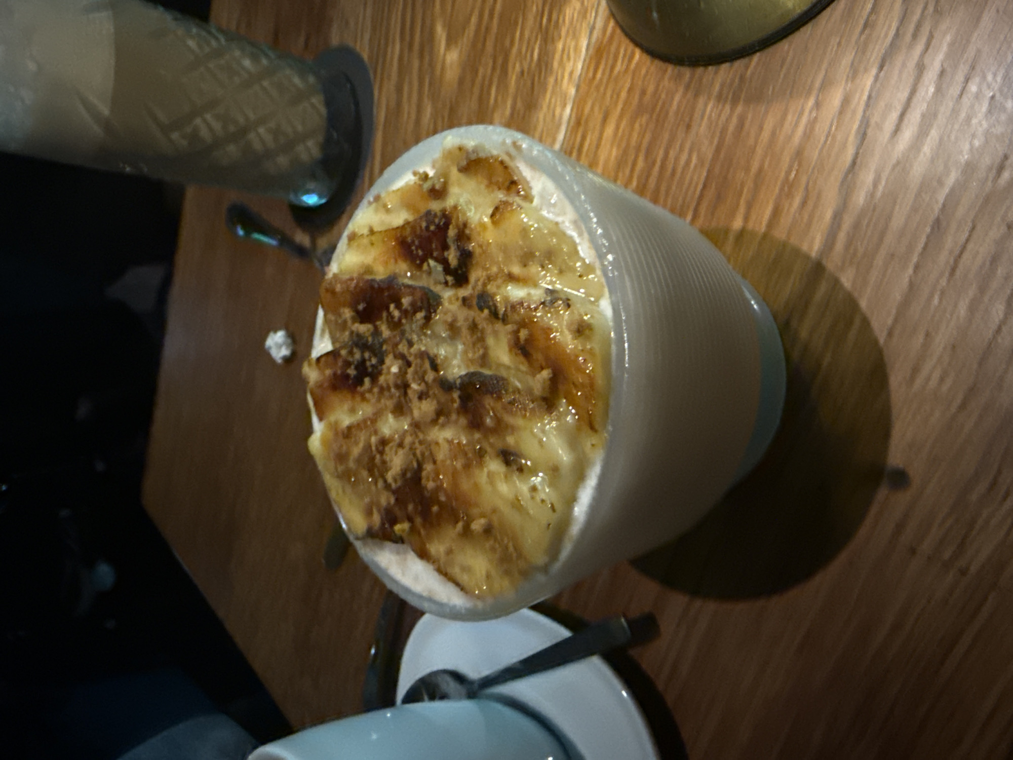 On a wooden table in what appears to be a restaurant, possibly in Mexico City, Mexico, based on cuisine, there are two cylindrical glasses/containers. One contains a white beverage, possibly a milkshake or smoothie, while the other holds a dessert topped with what appears to be caramelized banana and a granular topping. A small spoon rests on a saucer beneath the dessert. The scene is viewed from above, slightly off-center. The rest of the scene is out of view, except the corner of a white mug in the lower corner of the image.