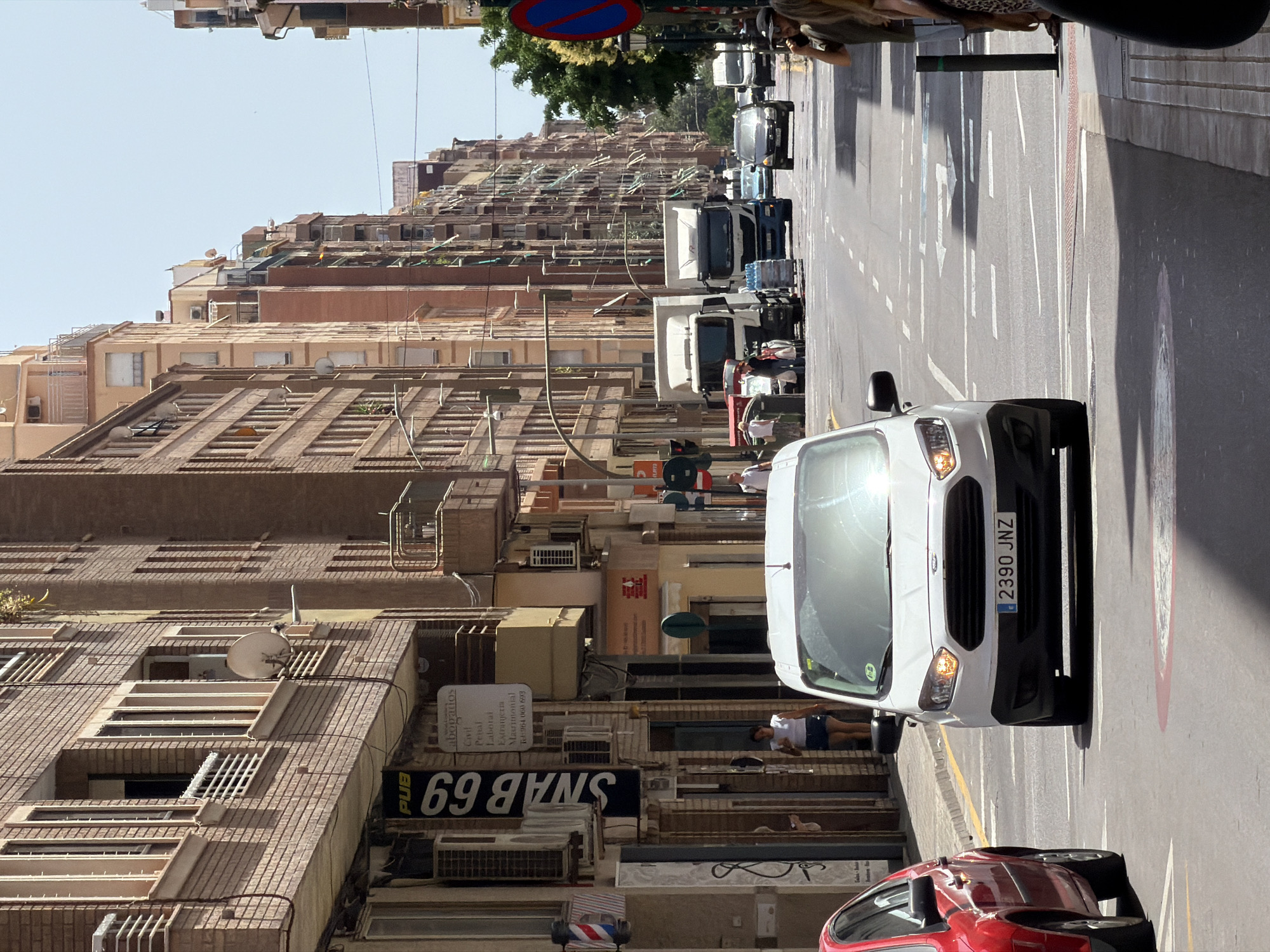 The image depicts a street scene in Castelló de la Plana, Spain, characterized by multi-story brick buildings lining both sides of a paved road. The architecture suggests a European urban environment with balconies, windows, and visible air conditioning units on the building facades. Sunlight casts shadows across the street, indicating a bright, sunny day.
In the foreground, a white van with the license plate "2390 JNZ" is prominent, positioned in the right lane of the road. To its left, the rear of a red car is visible. The road markings include lane dividers and directional arrows painted on the asphalt. Several other vehicles are present further down the street, including trucks, a dark-colored car, and another van. Pedestrians can be observed on the sidewalks and near some of the buildings, including a woman in a white top and dark shorts standing on the sidewalk to the left of the white van, and a man in a dark t-shirt talking on a phone on the right side of the road. A sign with "PUB SNAB 69" is affixed to one of the buildings. Traffic signals are visible, indicating a controlled intersection. The overall scene conveys a typical daytime urban street environment in Spain.
