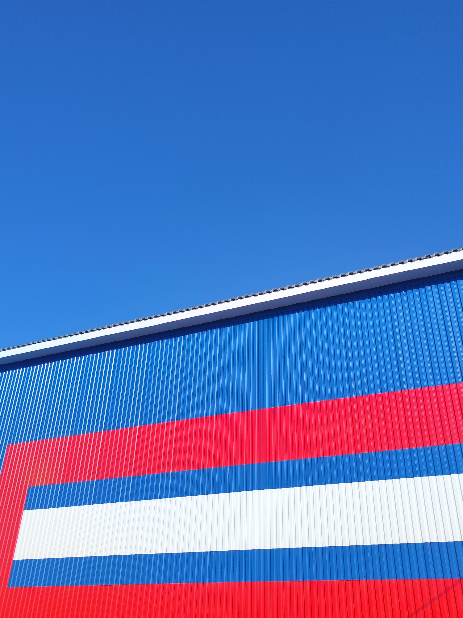 The image shows the side of a building painted in the colors of the Russian flag. The building has a corrugated metal exterior, painted blue at the top, followed by red, white, blue, and red stripes. The sky above the building is clear and blue. The location is Unknown, Unknown.