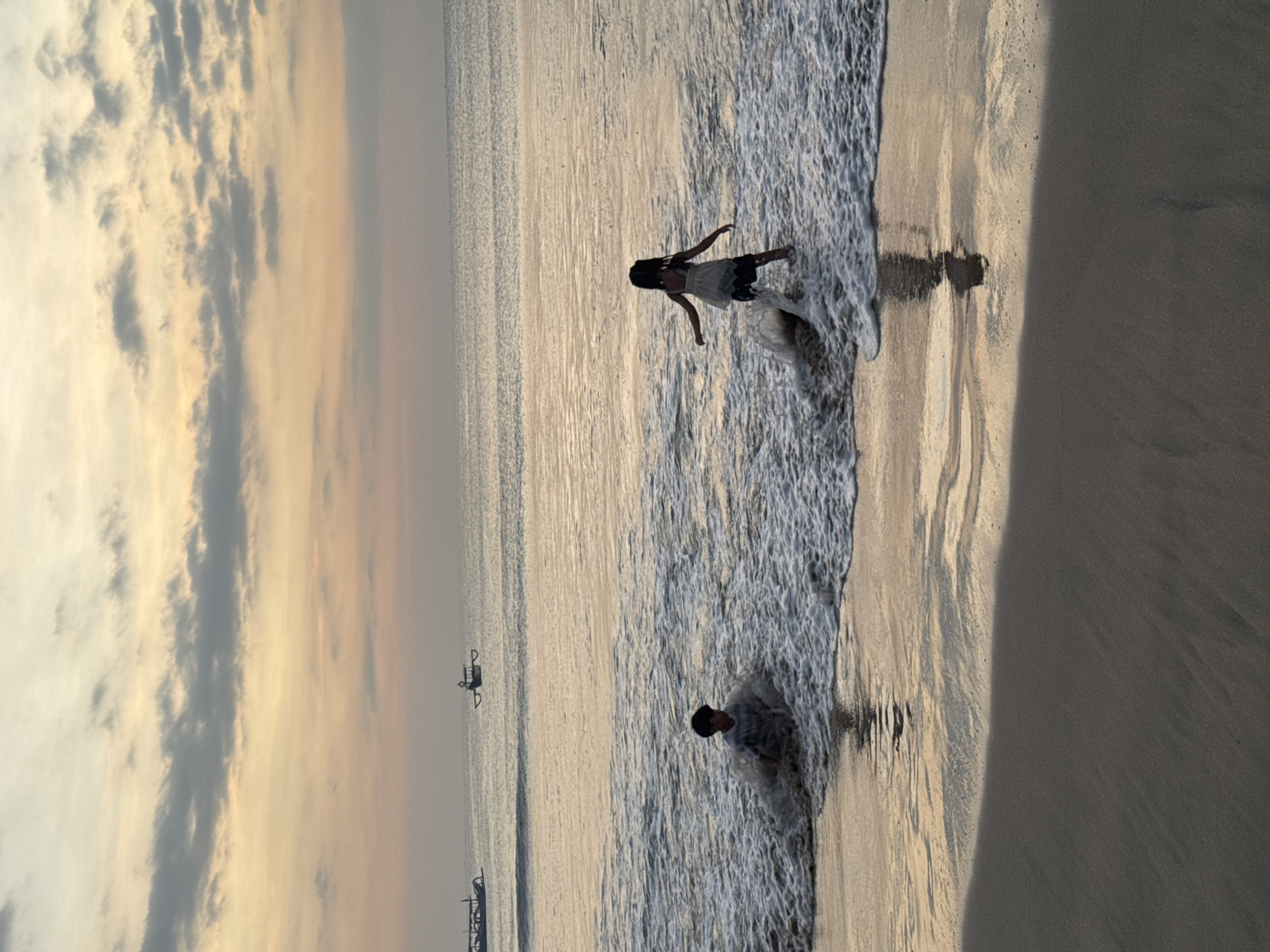 Two children, a boy and a girl, are playing in the gentle waves of the ocean at sunset on a beach in Kuta Selatan, Indonesia. The boy is seated in the water, with waves crashing around him, and he appears to be splashing. The girl is standing in the water, facing away from the camera, and her arms are outstretched as if she is about to move or interact with the water. The sky above is filled with soft, diffused light, indicating the sun is setting, with streaks of orange, pink, and gray clouds. Several traditional Indonesian fishing boats, known as jukungs, are visible in the distance on the calm sea. The wet sand of the beach in the foreground reflects the warm colors of the sky.
