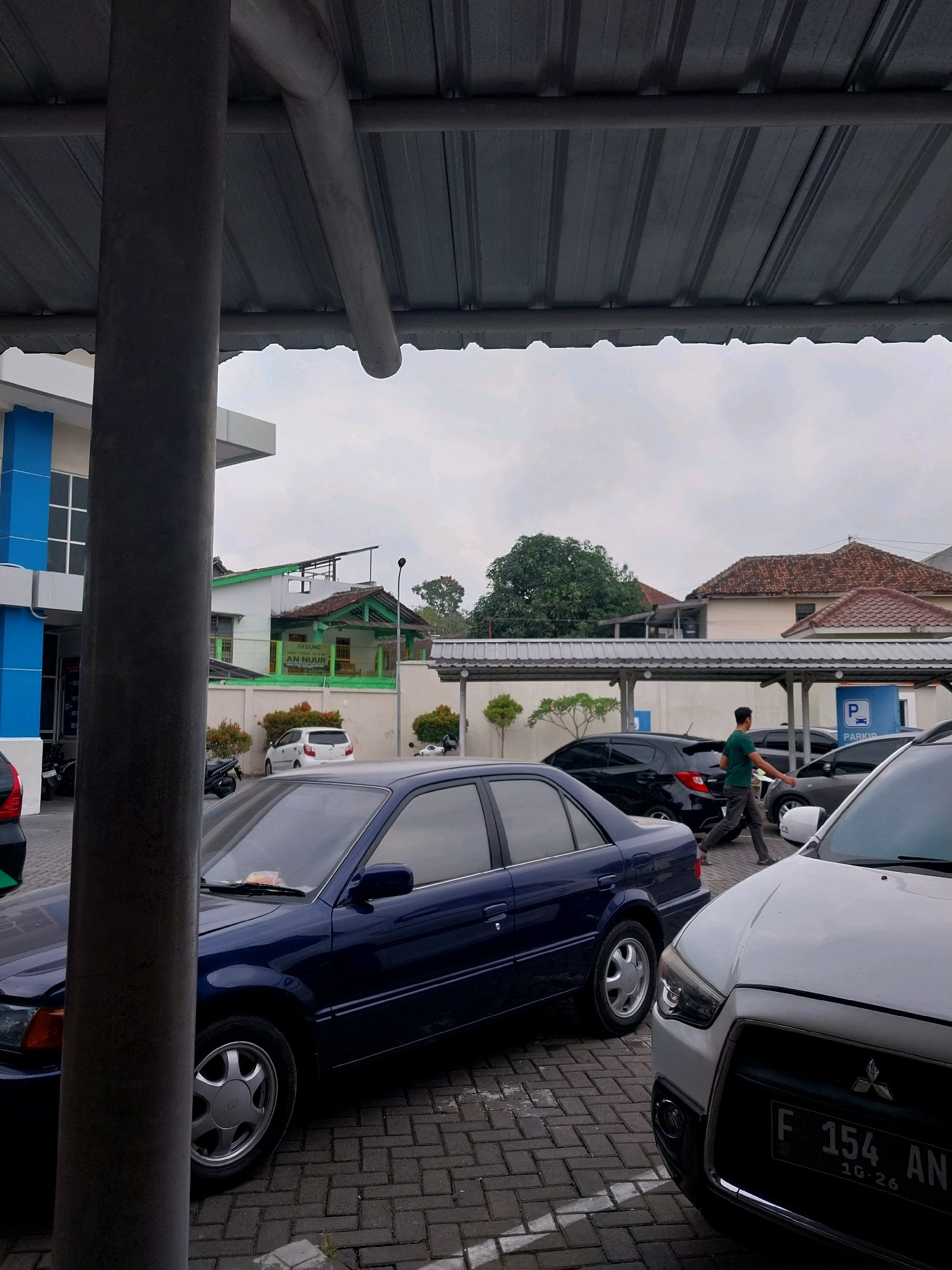 The image depicts an outdoor parking lot in Sleman, Indonesia, under a corrugated metal canopy supported by large, dark gray cylindrical pillars. In the foreground, a dark blue sedan is parked on a brick-patterned paved surface. To the right, the front of a white SUV is visible, displaying a license plate with the characters "F 154 AN" and "10.26". A man wearing a green t-shirt and dark pants is walking away from the viewer towards the right side of the frame, passing between parked vehicles. Behind the blue sedan, a black hatchback is parked. Further in the background, a white sedan is parked. Beyond the parking area, buildings with green trim and tiled roofs are visible, along with trees and an overcast sky. The overall scene suggests a typical day at a public parking facility.