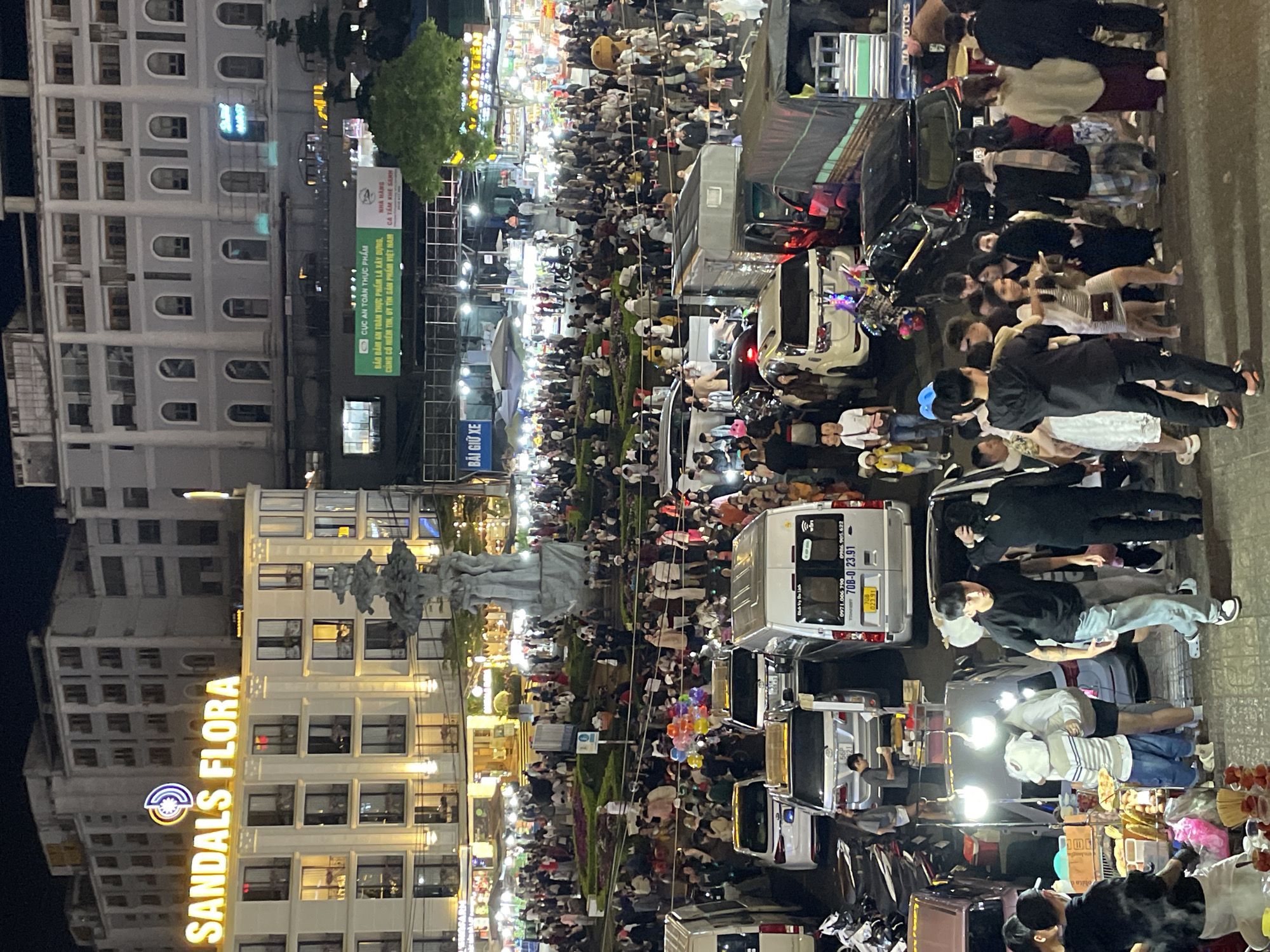 This is a nighttime aerial view of a busy street in Da Lat, Vietnam. A prominent building in the background displays a large illuminated sign that reads "SANDALS FLORA". The street is crowded with people, many of whom appear to be shoppers or tourists. Numerous vehicles, including vans, cars, and motorcycles, are present, suggesting a bustling commercial or entertainment district. The scene is illuminated by streetlights and the lights from buildings and stalls, creating a vibrant atmosphere. There is a large stone sculpture in the central area of the street, which seems to be a landmark. The ground is paved, and there are indications of food stalls or vendors selling goods to the crowds. The overall impression is one of a lively and active urban environment at night.
