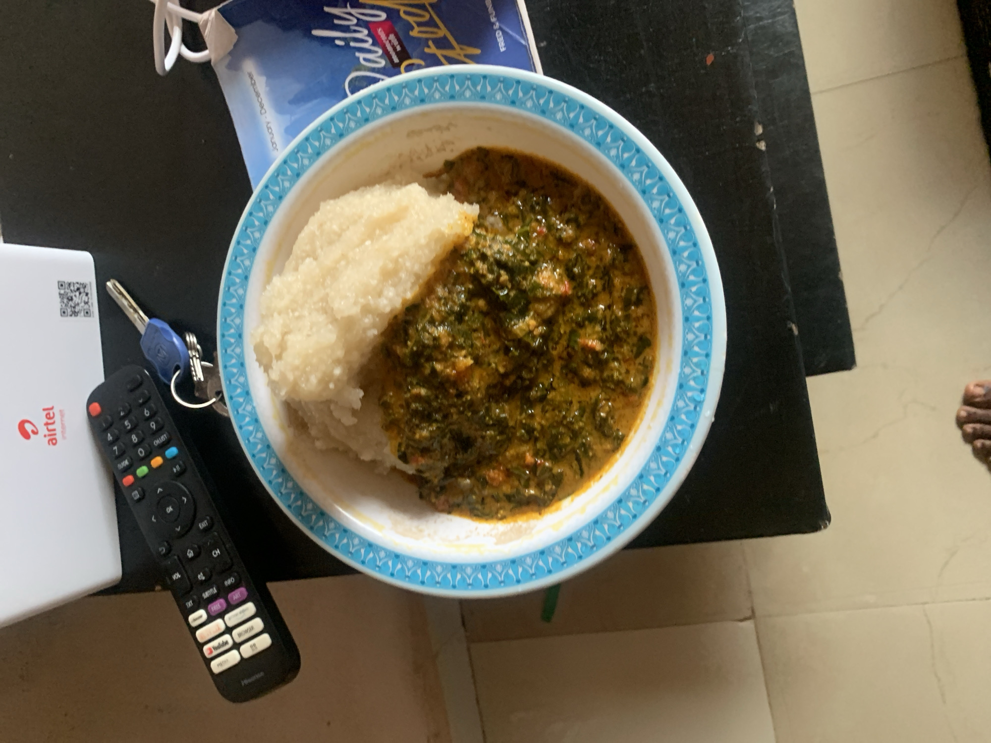In Sabon Tasha, Nigeria, a remote control and a bowl of food are placed on a dark surface. The remote, black with multicolored buttons, rests to the left of the bowl. The bowl itself is white with a blue decorative rim and contains a portion of light-colored food, likely a starch like fufu or ugali, topped with a vibrant green vegetable stew. To the right of the bowl, a blue and white magazine titled "Daily Food" is visible. The scene is illuminated by overhead lighting, casting soft shadows. The background shows a white Airtel internet router. The floor in the foreground is tiled with a light beige color. A partial view of a person's feet is visible at the bottom of the frame.