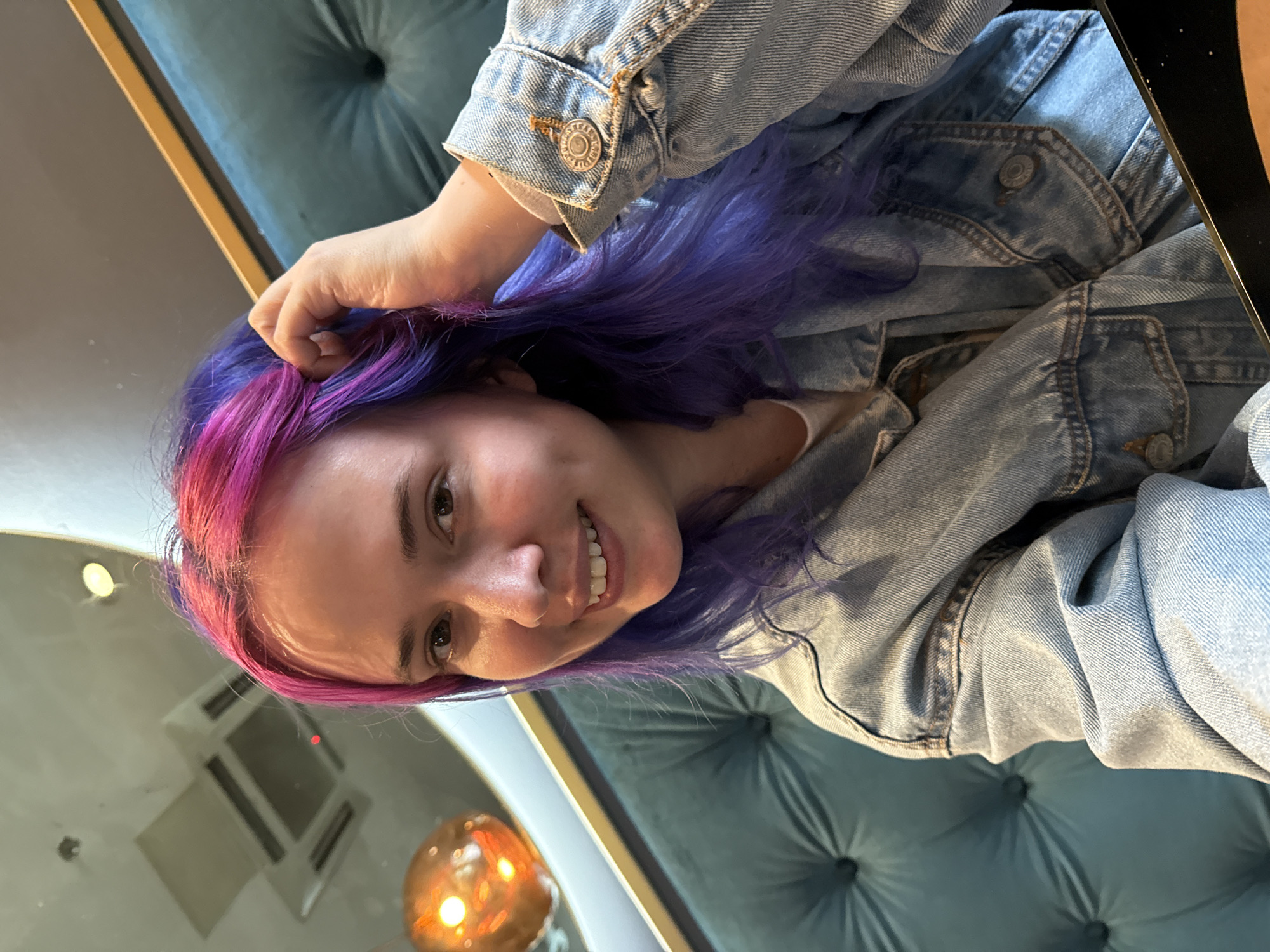 A young woman with dyed pink and purple hair is smiling at the camera. She is wearing a light-blue denim jacket. She is either sitting on or standing near a tufted blue-green upholstered object, possibly a chair or sofa. In the background, a room with a mirror and a light fixture is visible. There is no information to confirm this photo was taken in Zagreb, Croatia.