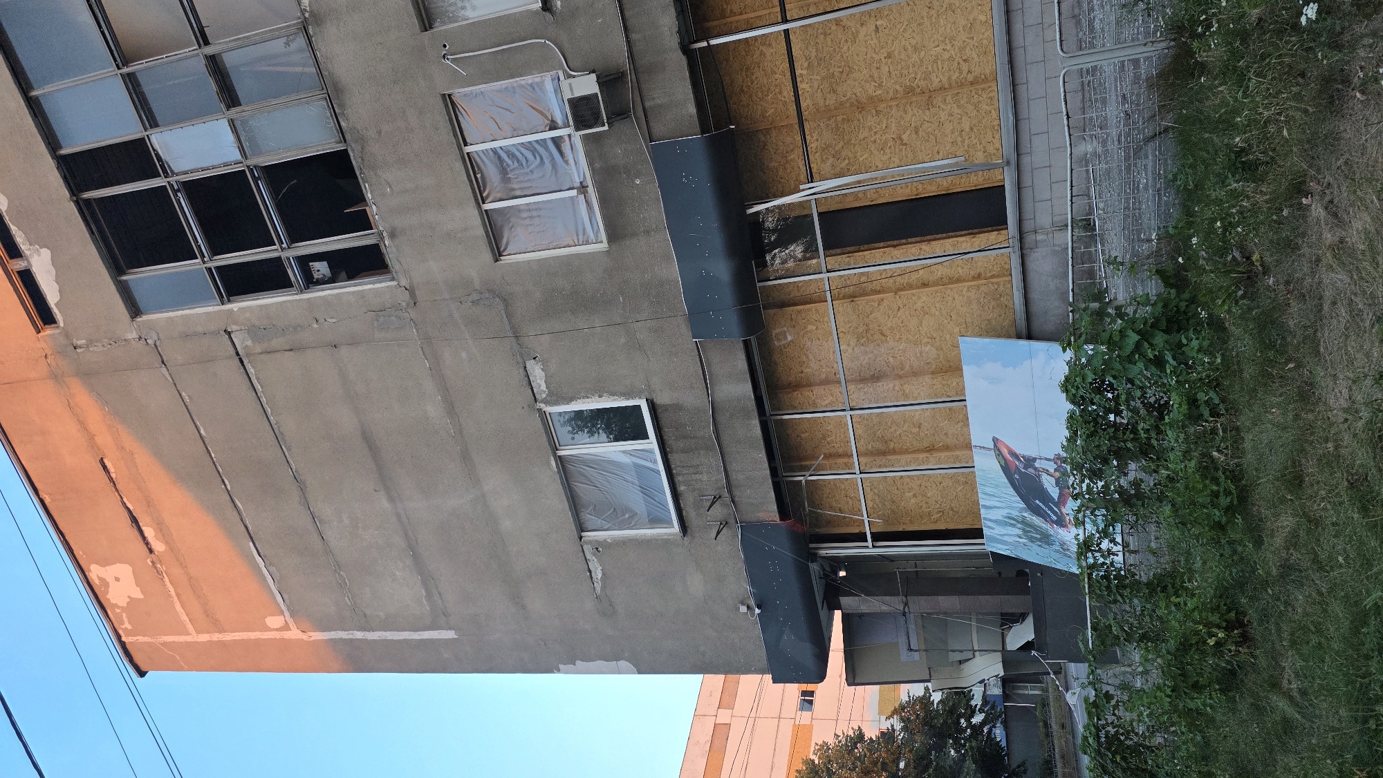 The image depicts the exterior of a multi-story building, likely in an urban setting. The building's facade is made of gray concrete, showing signs of wear and tear, including cracks and peeling paint. The upper floors feature several windows; some are intact, while others are broken or boarded up with plywood. One section of the building is illuminated by warm sunlight, casting a shadow across the concrete.
The ground floor appears to have been a commercial space, with large windows that are now entirely covered with plywood. A dark, horizontal awning stretches across this level. In front of the building, there is a low metal fence bordering a grassy area. A large, colorful advertisement is propped against the fence, displaying an image of a person on a jet ski. Greenery, including bushes and grass, grows in front of the fence.
In the background, another building is visible, appearing to be a modern, multi-story structure with orange and white elements. The sky is a clear blue, with visible utility wires running across the top of the frame. The overall scene suggests a potentially abandoned or under-renovation commercial property. The presence of the advertisement and the contrast between the weathered main building and the newer structure in the background might indicate an area undergoing change or with a mix of old and new developments. The weather appears clear and sunny.