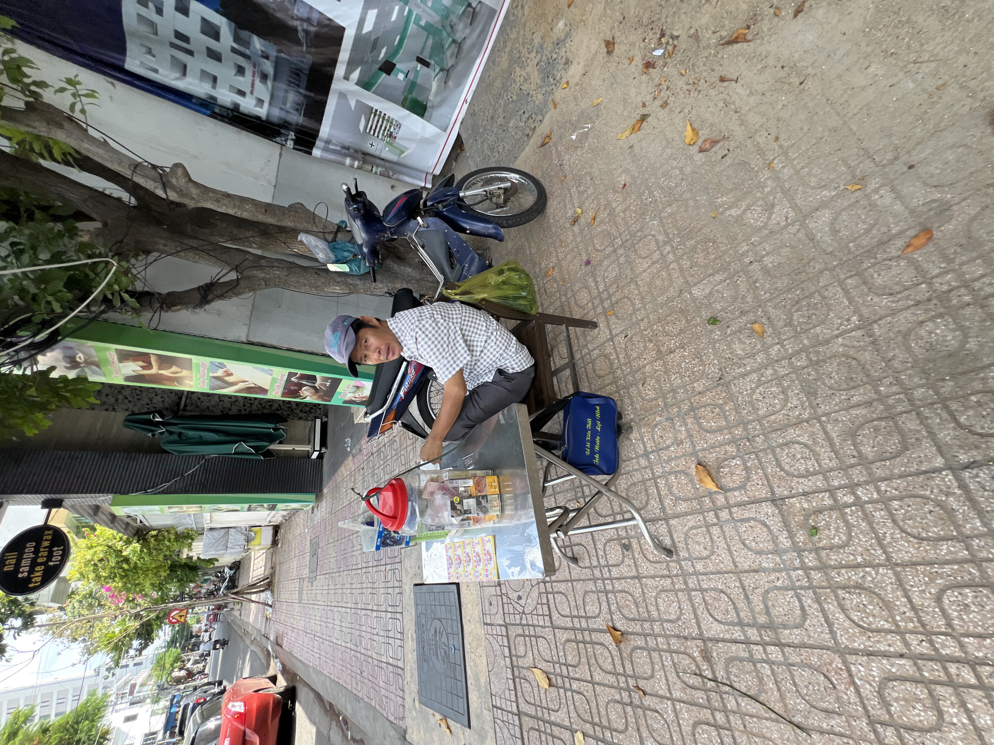 A middle-aged Vietnamese man wearing a grey baseball cap and a checkered short-sleeved shirt sits on a stool at a metal table on a sidewalk in Nha Trang, Vietnam. The table is laden with items, including a large plastic jar filled with what appears to be drinks or candy, several packs of cigarettes, and other small packaged goods. A blue tote bag with Vietnamese writing is placed on the ground beneath the table. A dark blue motorcycle is parked behind the table, and a green plastic bag hangs from the back of the stool. The sidewalk is paved with patterned tiles and is lined with trees and a building with a sign that reads "nail shampoo take earwax foot." In the background, a red car is parked on the street. The scene suggests a small, informal vendor operating on the street.