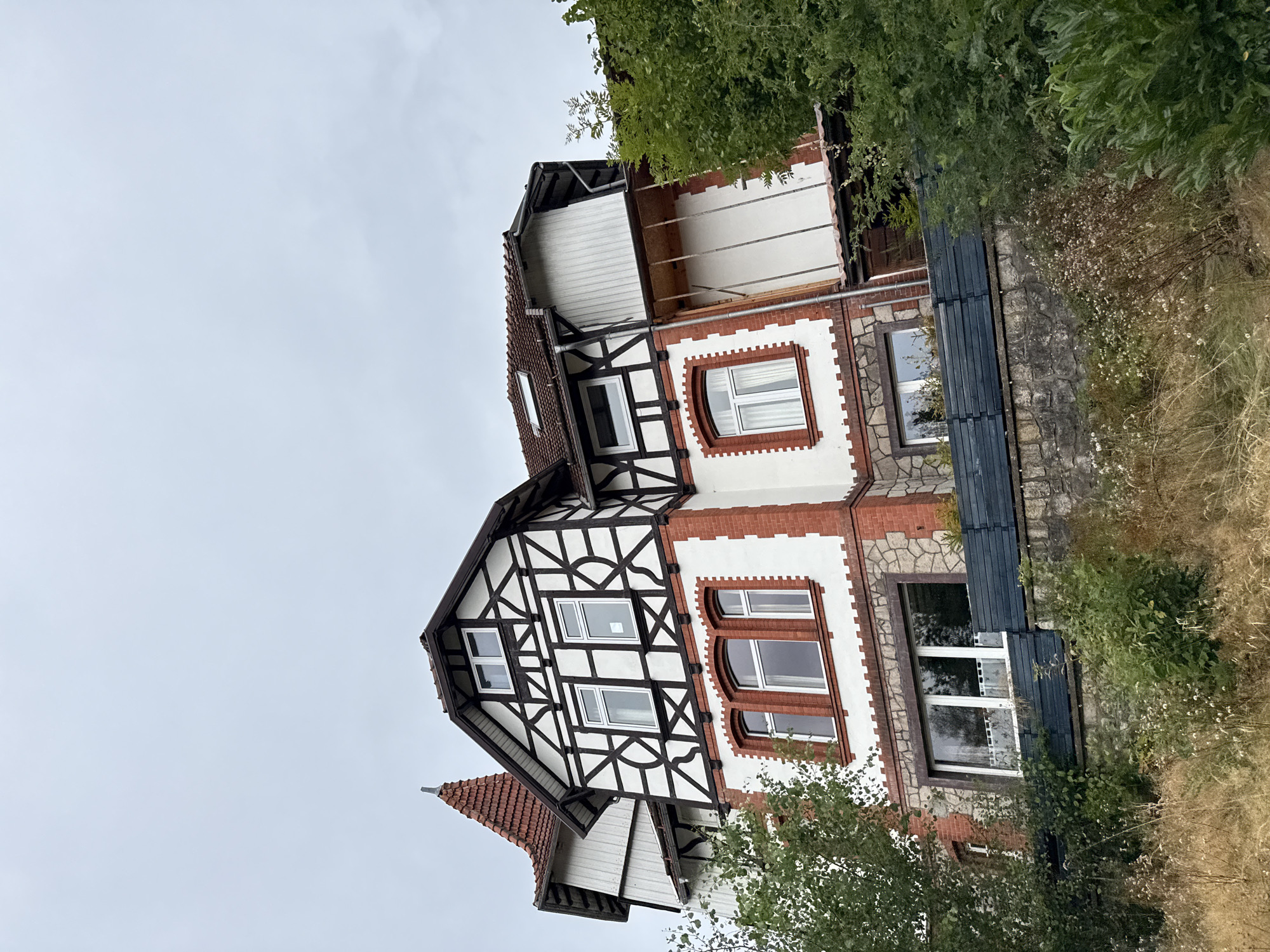 A half-timbered house with a red tiled roof and a pointed turret stands on a hillside under a cloudy sky. The house has white stucco walls with dark brown timber framing and red brick accents. Several windows with white frames are visible, some with curtains. A dark blue fence runs along the front of the property, with a stone wall below it. The foreground is filled with dry grasses and green bushes. The house is located in Bad Sachsa, Germany.