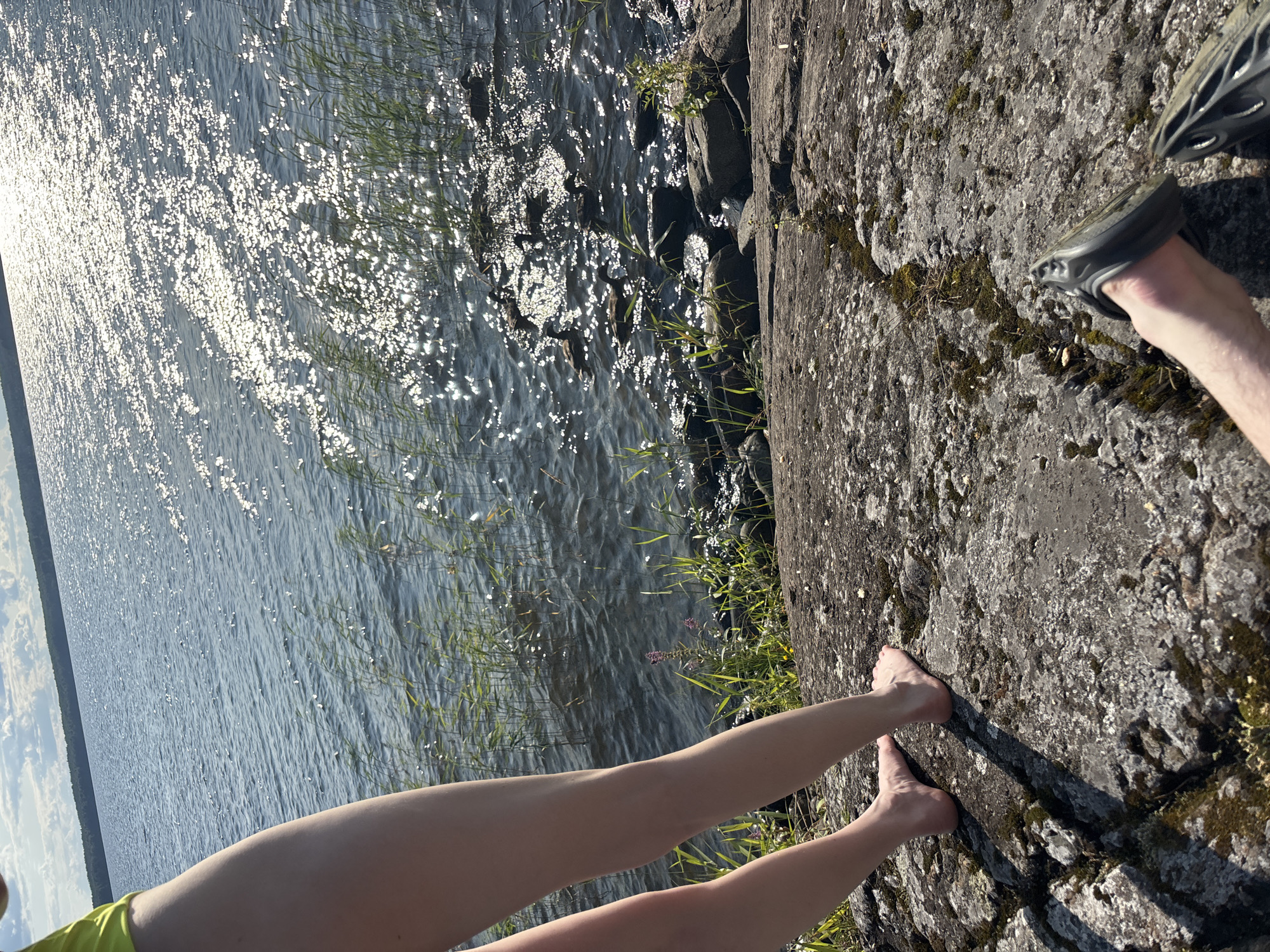 The image depicts a sunny day by a body of water, likely a lake. In the foreground, a person's bare legs and feet are visible, standing on a large, uneven rock formation. The rock is weathered and has patches of moss. To the right, another person's foot is clad in a dark sandal, resting on a similar rock surface.
The water is calm with ripples reflecting the bright sunlight, creating a shimmering effect. Reeds and grasses grow along the edge of the water, interspersed with dark rocks submerged at the shoreline. In the distance, a tree line forms the horizon, under a sky with scattered white clouds.
The location is identified as Shuiskaya Chupa, Russia, suggesting a natural, outdoor setting typical of a lakeside recreational area. The overall scene conveys a sense of leisure and enjoyment of nature.