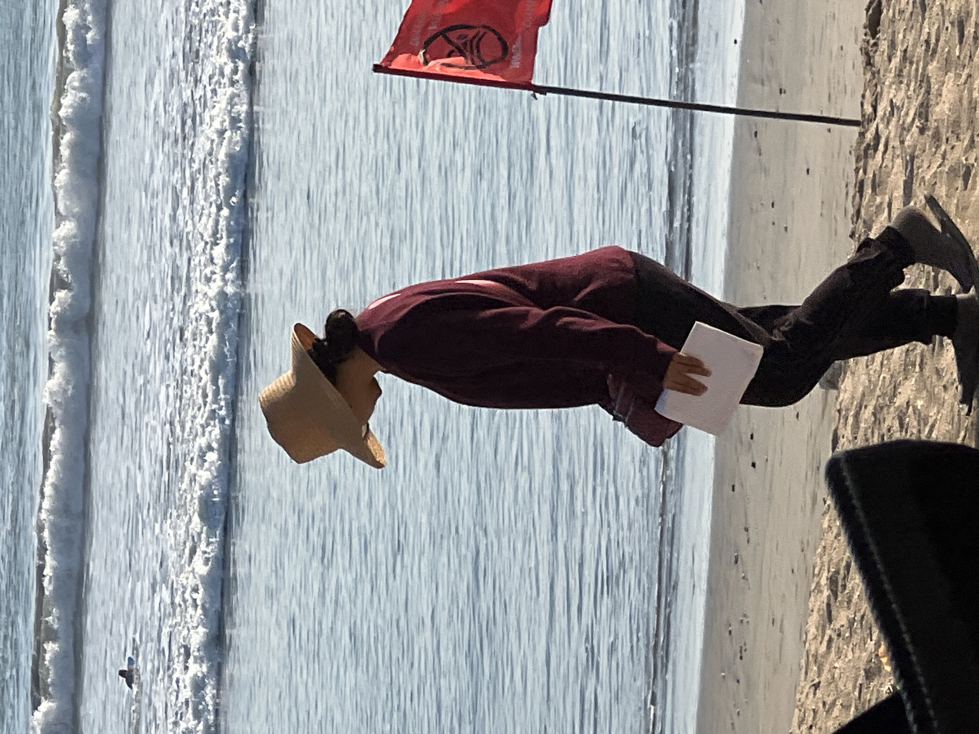 The image depicts a person wearing a straw hat, a maroon sweater, and dark pants, walking on a sandy beach. The person is holding a white piece of paper. To the right of the person, a red flag with a graphic symbol is planted in the sand on a pole. The ocean with breaking waves is visible in the background, and a surfer on a blue surfboard can be seen in the distance. The lighting suggests it is daytime, possibly late afternoon due to the long shadows. The scene appears to be a public beach.