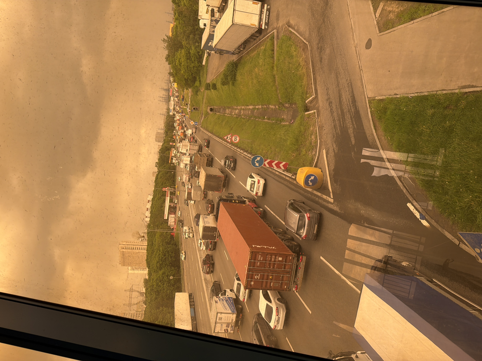 The image depicts heavy traffic congestion on a highway in Moscow, Russia, during a sandstorm or other atmospheric event causing significantly reduced visibility. The view is from an elevated position, likely a high-rise building, looking down onto the multi-lane highway. Multiple trucks and cars are visible, mostly stationary or moving slowly due to the gridlock. The sky is a hazy, orange-brown color. There are patches of green vegetation alongside the road. Several buildings are seen in the distance and obscured by dust. A red shipping container and numerous other vehicles are clearly visible in the closer view.