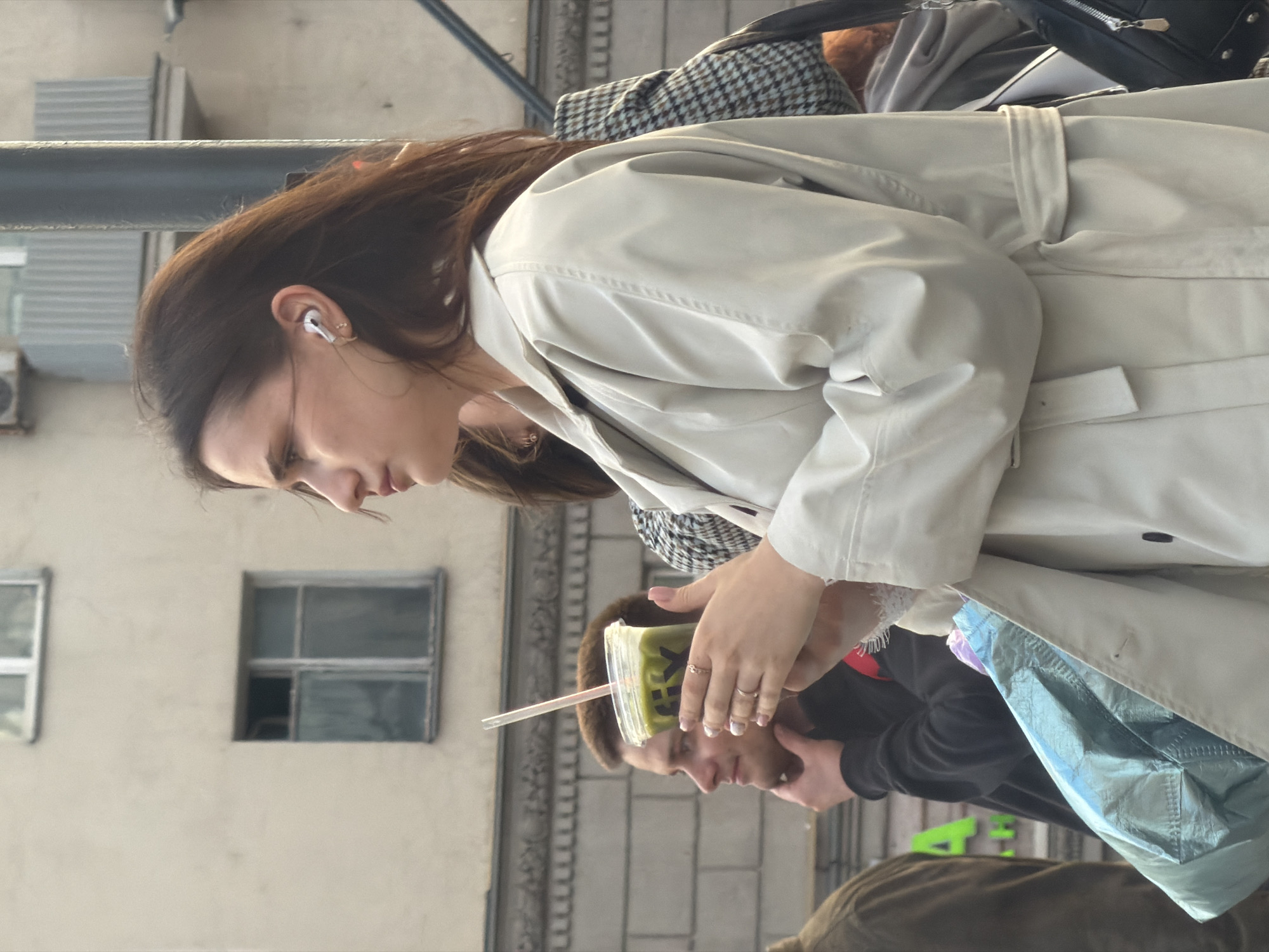 A woman with long brown hair and wearing an off-white trench coat stands outdoors, holding a clear plastic cup with a green beverage and a straw. She has a white wireless earbud in her left ear. A man is seen just below the cup, holding the cup steady with his hand. Behind the woman are building facades with windows and architectural details. The scene is on the street in the city of Moscow, Russia.