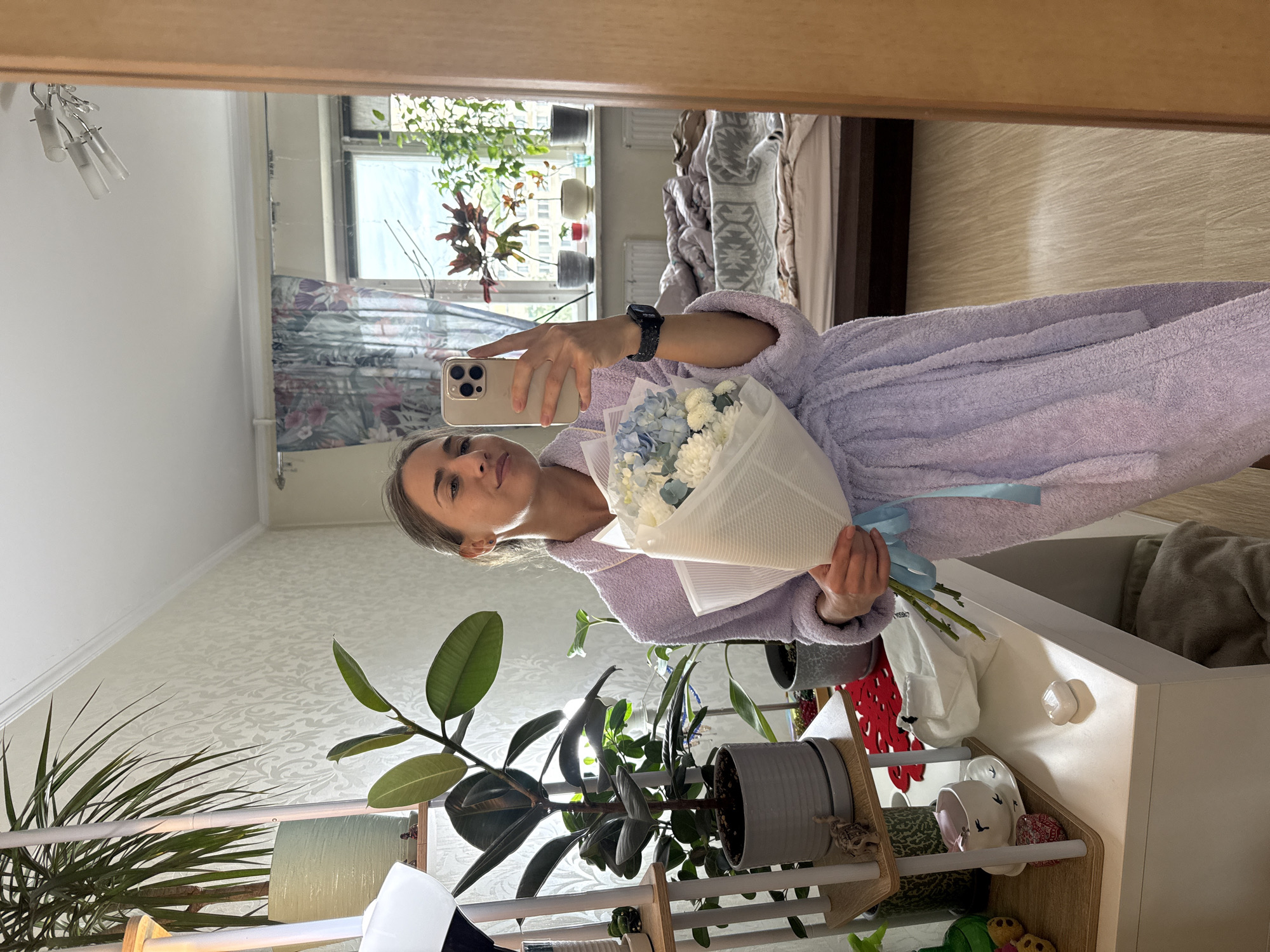 A woman in a light purple robe is taking a selfie in a mirror. She is holding a bouquet of white and blue flowers wrapped in white paper with a light blue ribbon. In her left hand, she holds a tan-colored smartphone. She is wearing a watch on her left wrist. The mirror reflects a room with a window, plants, and a bed. In the foreground, there are several potted plants on a shelf. The wallpaper is off-white with a subtle pattern. The location is Unknown, Russia.