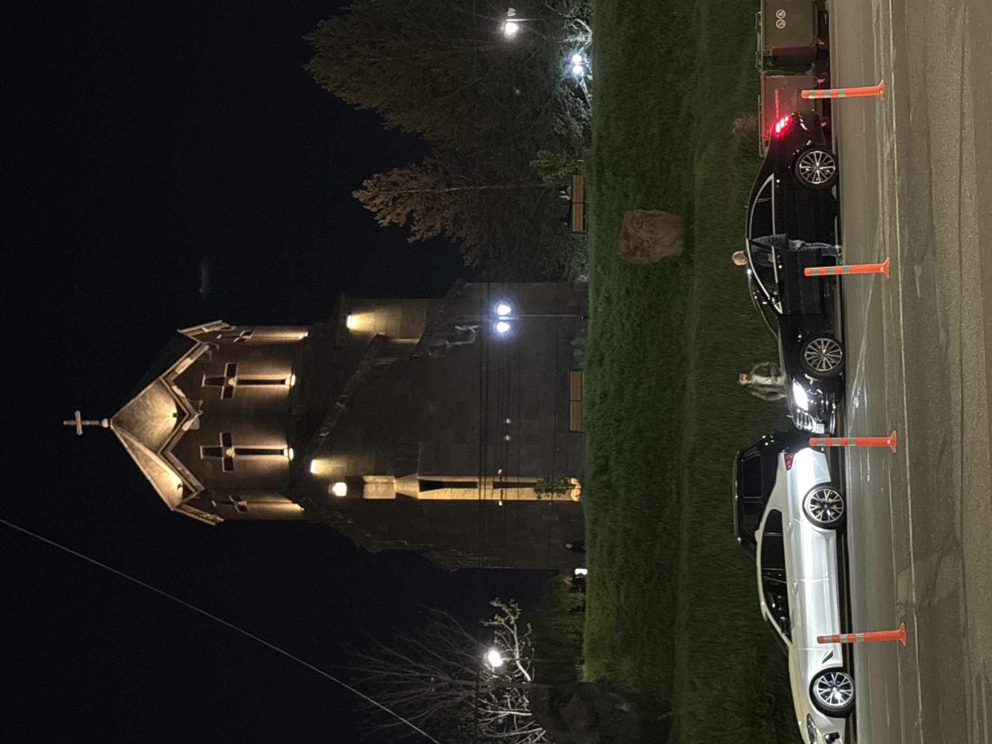 The image displays a night-time scene in Yerevan, Armenia, featuring a prominent church, a park area, and a street with parked cars and people. The overall setting is dark, illuminated by artificial lights.
On the left side of the frame, a stone church with distinct Armenian architectural characteristics stands illuminated against the dark sky. Its multi-faceted dome is topped with a cross, and several smaller crosses are integrated into the building's facade, highlighted by external lighting.
Adjacent to the church is a grassy area dotted with trees and several streetlights, casting light and shadows. Faintly visible park benches are nestled among the trees. A large, naturally shaped stone or sculpture rests on the grass closer to the foreground.
To the right, an asphalt street is visible. Two modern cars are parked along the curb, separated by orange traffic cones. The rear vehicle is black, with its red taillights illuminated, and a person is standing by its open driver's side door. The front car is silver or white. Another person is seen standing between the two cars, facing towards the silver vehicle. Behind the black car, green trash bins are placed on the sidewalk, featuring indistinct symbols, possibly recycling logos. The sky is dark and clear, suggesting a calm night. No clear readable text is discernible in the image.