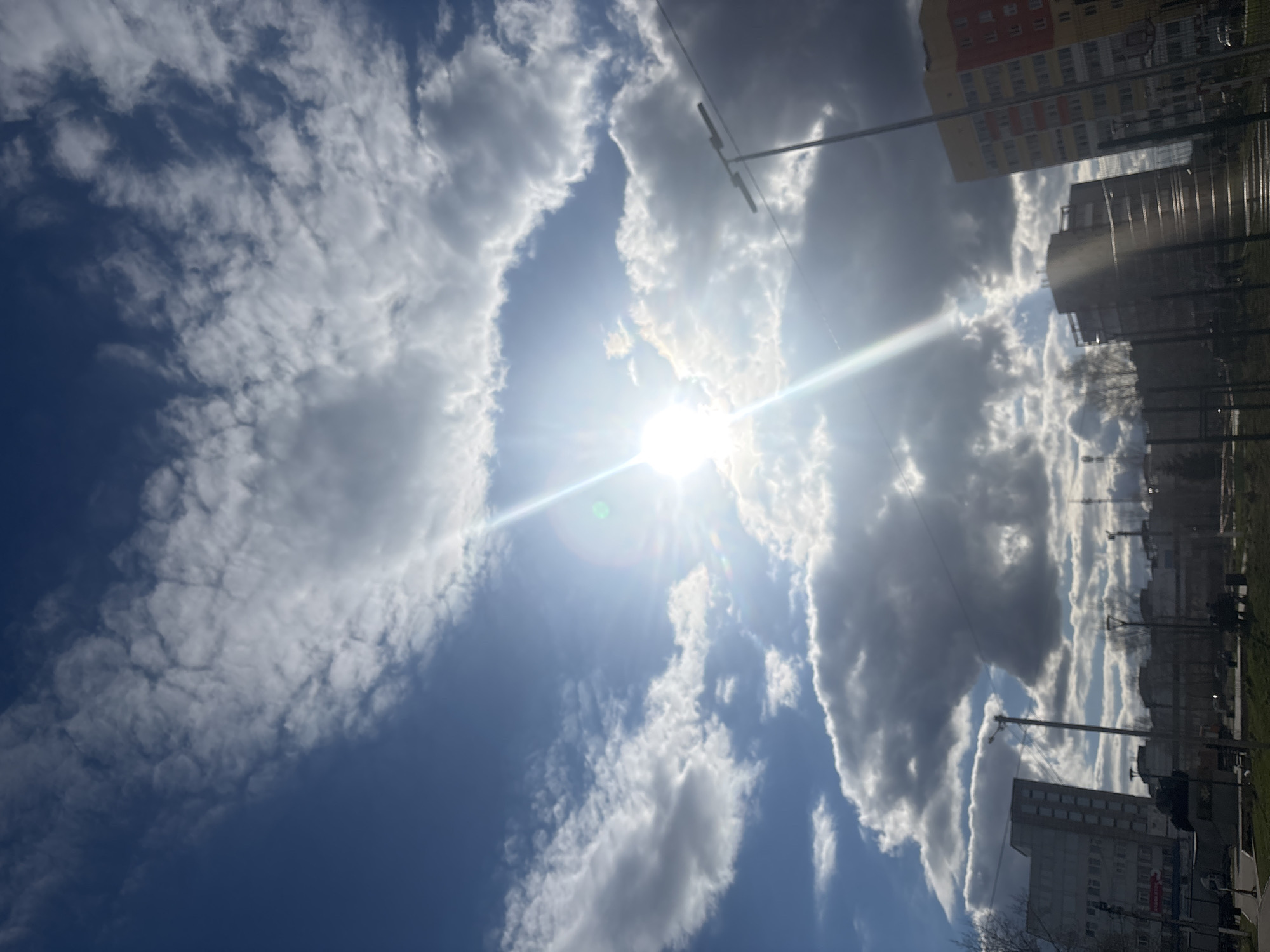 The image captures a dynamic sky over an urban landscape in Chaikovsky, Russia, viewed on a bright, partly cloudy day. The sun, a central and dominant feature, shines intensely from a gap in the clouds, casting a strong glare and producing distinct sunbeams and a lens flare.
The sky is a vibrant blue, featuring a diverse array of clouds. On the left side, there are wispy, scattered white clouds, suggesting higher altitudes or thinner formations. Towards the right and closer to the sun, the clouds become denser and more voluminous, ranging from bright white to darker grey, indicating significant cumulus or stratocumulus formations.
In the lower portion of the scene, several multi-story buildings characteristic of an urban or suburban environment are visible. One building stands out with distinct red and yellow painted sections, suggesting a residential block. Nearby are other structures, including a grey-toned building that appears industrial or commercial, partially obscured by wires and shadows. Utility poles or streetlights with overhead wires are prominent against the bright sky and buildings, adding to the urban feel. Fences or barriers are also discernible at ground level.
Hints of vegetation, such as bare tree branches and patches of green grass, are present along the base of the buildings, suggesting either early spring or late autumn. The overall impression is a clear, yet dynamically cloudy day, highlighting the contrast between the intense sunlight and the varied cloudscape above the city. No legible text is visible on any of the structures or signs.