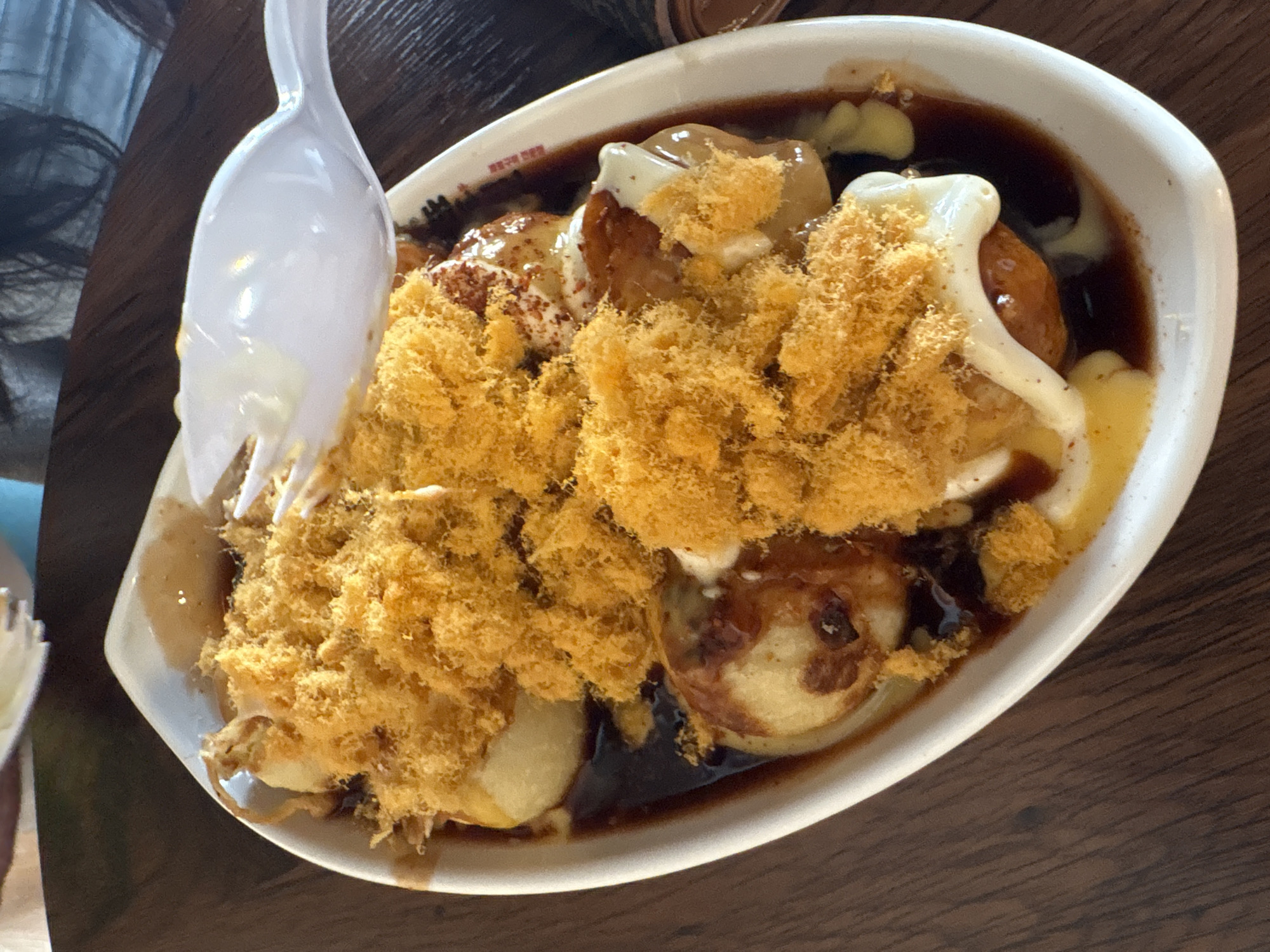 A white oval dish filled with several golden-brown, round food items, generously topped with shredded, golden-brown meat floss and drizzled with dark sauce and white sauce. A white plastic spoon is positioned over the dish, with some food clinging to its tines and surface. The dish rests on a dark wooden table. In the upper background, a blurred individual is partially visible, suggesting a dining environment. The scene appears to be in Davao City, Philippines, as indicated by the visible text on the dish's rim which reads "BISAG KISA ANG KASANGLAN" and "DAVAO CITY," the latter appearing in red ink.