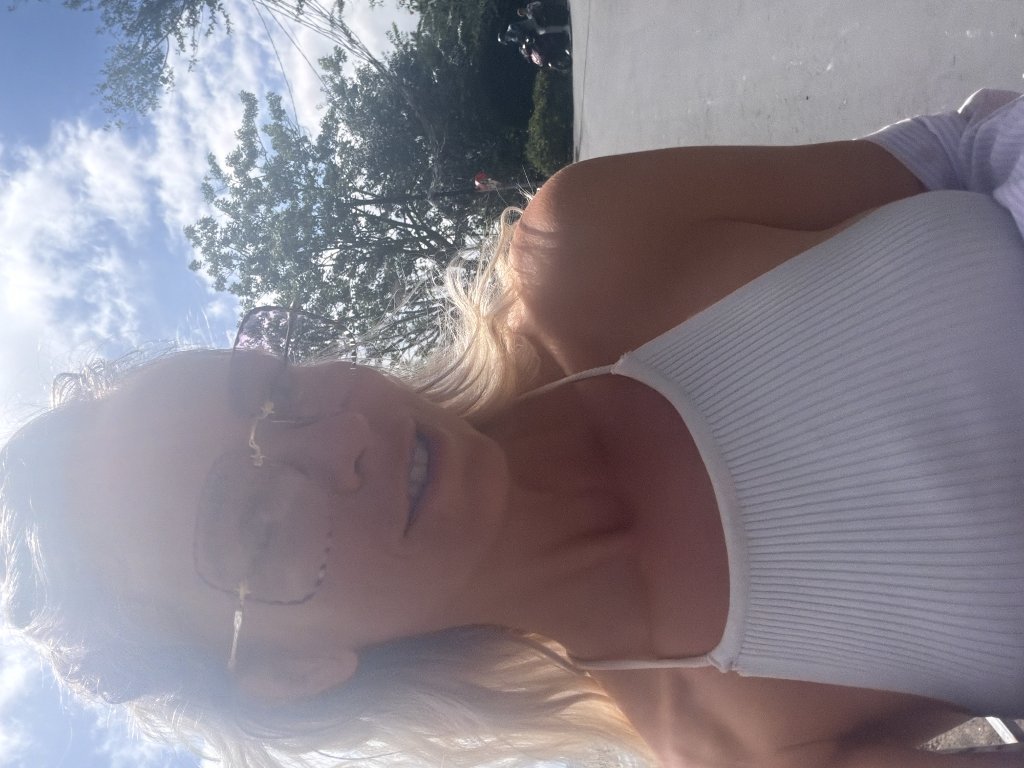 A woman with blonde hair, wearing oversized pink-tinted sunglasses and a white ribbed tank top, is captured in a close-up selfie. The bright sunlight causes lens flare, obscuring some details of her face. Her left shoulder and upper chest are visible, along with the strap of her tank top. Behind her, the sky is blue with scattered clouds. Trees with green foliage are also visible. In the background, there's a glimpse of a road or paved area where two people are riding on scooters. The overall scene suggests an outdoor setting, possibly in a moderately populated area within the city of Xuân Thành, Vietnam.