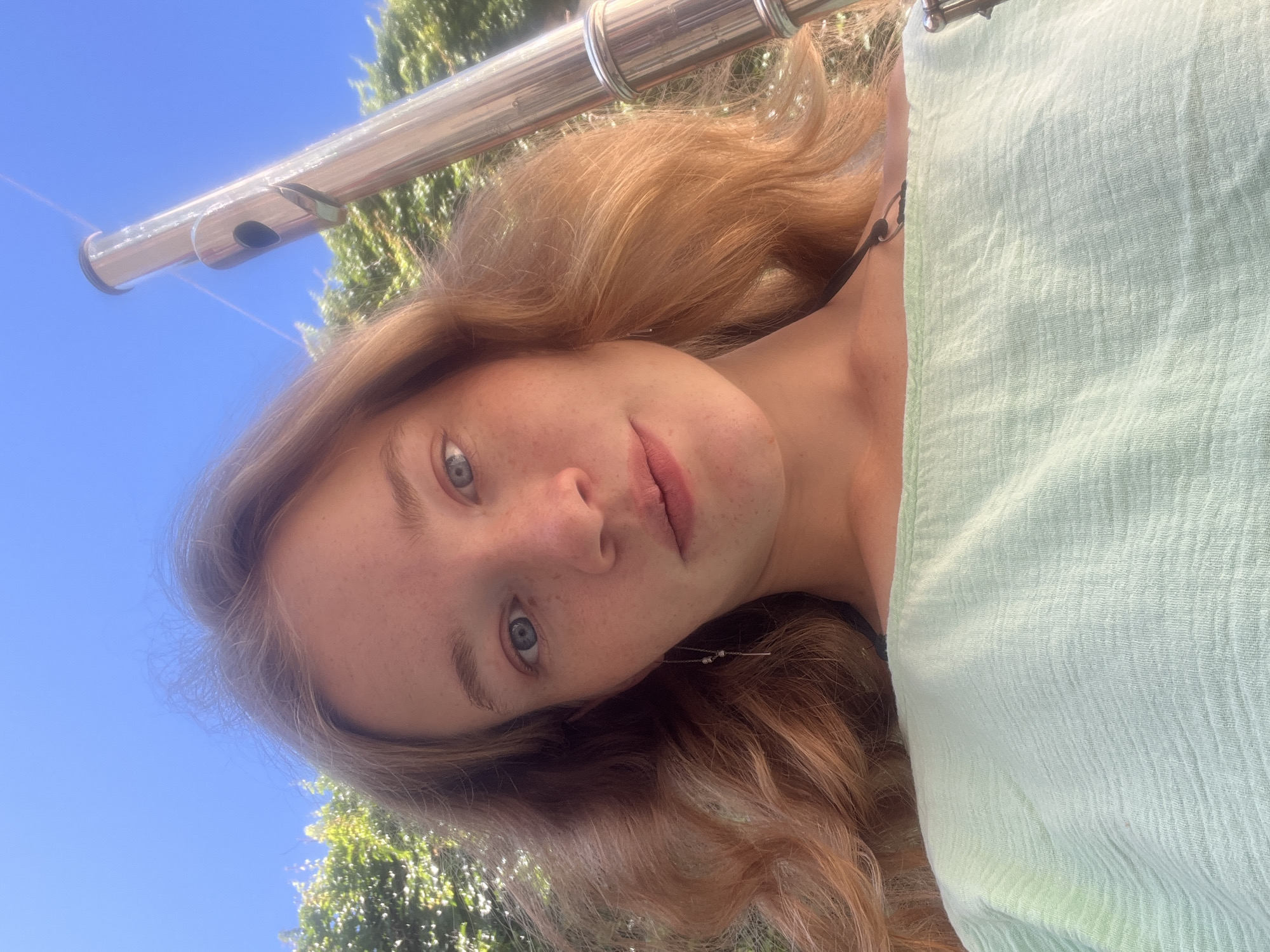 A young, fair-skinned woman with red-blonde wavy hair, light-blue eyes, and freckles is posing in what appears to be an outdoor setting. She wears subtle makeup with a muted lip color. Part of a metal flute is positioned above her head. She wears a light green textured garment on her right side. The background shows a bright blue sky with green foliage from trees partially visible behind the flute and her hair. While the scene is outside, it is impossible to identify any specific objects that would definitively identify the location as Budva, Montenegro.