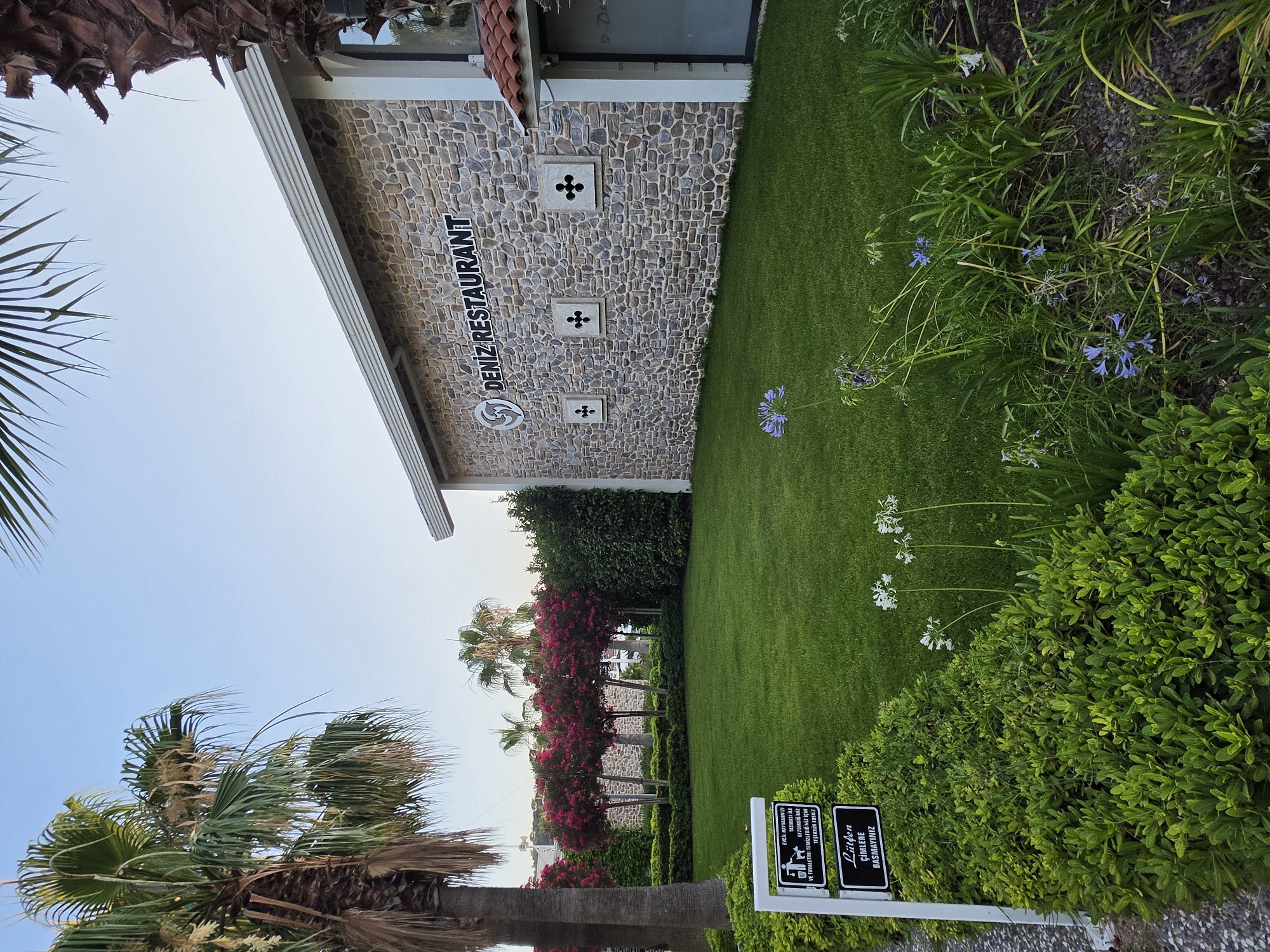 This is a sideways outdoor photograph featuring the exterior of a restaurant called "DENİZ RESTAURANT". The building is constructed with stone and has a red-tiled roof. Green grass lawns surround the establishment, and there are blooming pink flowers on bushes and trees. Palm trees are also visible, suggesting a warm climate.
The image appears to be taken during daylight, with a clear blue sky. There are no people visible in the photograph. Two signs are present near the bushes. One sign, written in Turkish, thanks visitors for walking their pets and cleaning up after them. The other sign, also in Turkish, requests people not to step on the grass. The overall atmosphere is serene and inviting, typical of a well-maintained outdoor dining area.