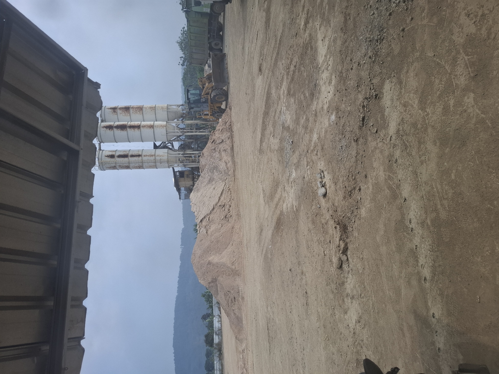 The image depicts an outdoor industrial scene, likely a construction or material processing site, under an overcast sky. In the foreground, a vast expanse of dusty, reddish-brown earth dominates the frame, showing tire tracks and scattered debris. To the left, a portion of a corrugated metal roof structure extends into the frame from above. In the mid-ground, a large pile of light brown sand or aggregate is visible. Behind the sandpile, three tall, cylindrical silos, rusted and weathered, stand prominently. To the right of the silos, a yellow front-end loader is positioned, its bucket lowered. Further to the right, a large green truck with a cargo bed is parked. The background features a line of trees and a gentle, rolling hillside or mountain range. The location is identified as Kutasirna, Indonesia.
