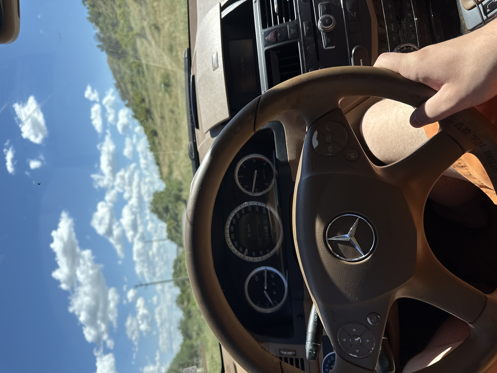 The image is taken from the driver's perspective inside a Mercedes-Benz car, likely in Sotirya, Bulgaria, on a sunny day. The focus is on the steering wheel, dashboard, and the view through the windshield. The steering wheel is a tan leather, with a Mercedes-Benz emblem in the center. To the left of the steering wheel is the turn signal lever, and on the right, a hand is visible gripping the wheel. The dashboard displays the car's instruments, including a speedometer showing 61 km/h, and other gauges. The car's interior appears to be well-maintained. Through the windshield, a road lined with green trees and rolling hills can be seen under a bright blue sky with scattered white clouds. The environment suggests a rural or semi-rural setting.