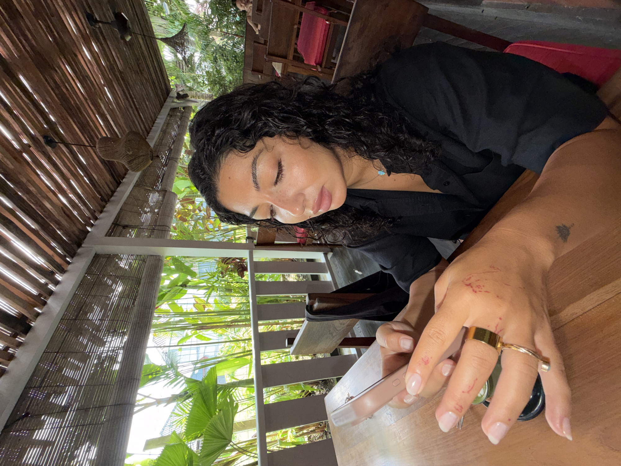 This image captures a woman resting in a bright, open-air tropical setting, likely in North Kuta, Indonesia. The subject, a woman with dark, curly hair, light brown skin, and closed eyes, appears to be relaxing or sleeping, with her head gently bowed and resting on a wooden table. She is dressed in a black collared shirt and wears a delicate light blue pendant necklace.
Her left hand, holding a light-colored smartphone, is prominent in the foreground. This hand features a wide gold ring on her middle finger and a twisted gold ring connecting her middle and index fingers. Her fingernails are manicured with a natural-looking finish. Notably, there are several small red marks, resembling scratches or insect bites, visible on the back of her fingers and hand. A dark tattoo is partially visible on her outer forearm.
The scene is an open-sided structure, possibly a restaurant or cafe, characterized by a slatted wooden ceiling and woven bamboo screens that act as walls, allowing light to filter through in distinct patterns. Beyond the structure, abundant lush green tropical foliage, including large palm leaves and bamboo, creates a vibrant natural backdrop, visible through a wooden balustrade. A red cushion on a wooden chair is faintly visible in the interior background. The strong, filtered light indicates it is daytime. No discernible text is visible.