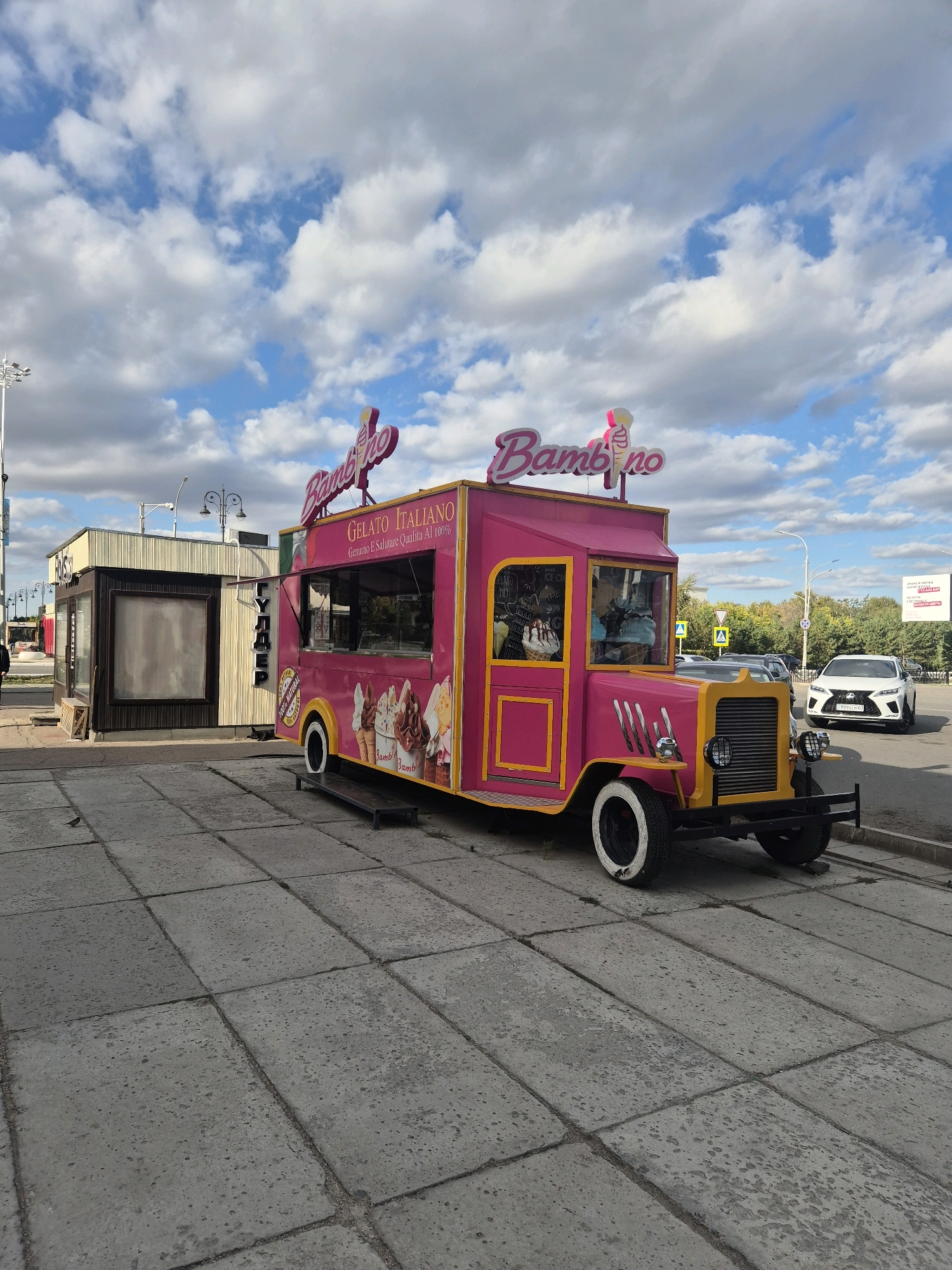 The image depicts an outdoor scene in the city of Kokshetau, Kazakhstan, likely on a sidewalk adjacent to a street. A vibrant pink and yellow gelato truck, branded with "Bambino" and featuring images of ice cream cones, is prominently positioned in the foreground. The truck is parked on a concrete tiled surface. To the left of the gelato truck is a smaller kiosk or stand with a dark brown facade and light-colored siding. In the background, there is a street with traffic, including a white Lexus SUV, and trees lining the roadside under a partly cloudy sky. Streetlights and traffic signs are also visible. The overall impression is a commercial area, possibly a public square or promenade, on a day with mixed cloud cover.
