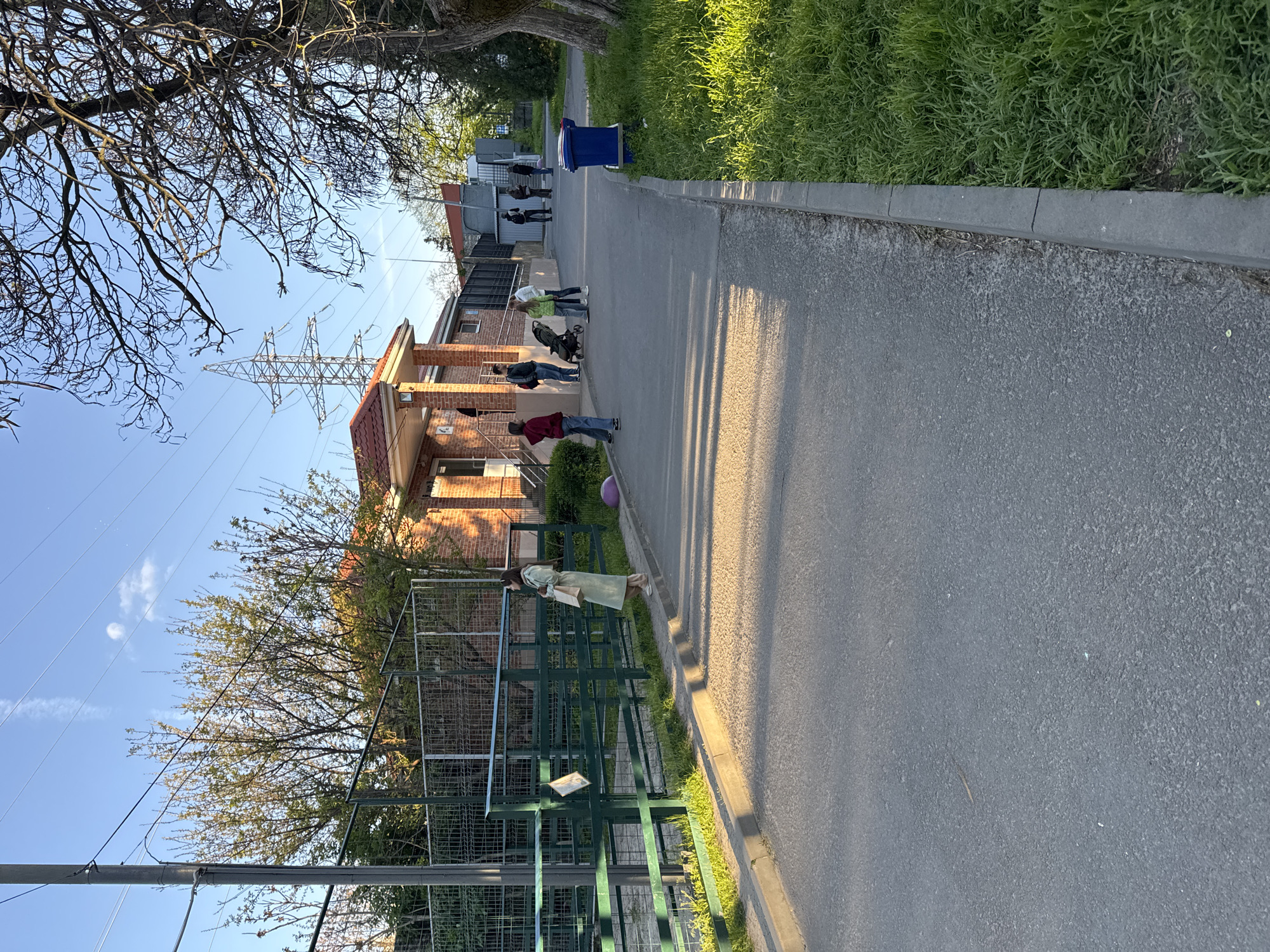 The image depicts a sidewalk in a residential area, possibly in Rostov-on-Don, Russia, though this location cannot be confirmed with certainty without additional evidence. A line of people is standing on the sidewalk, appearing to be waiting or queuing. A building with a brick facade and a red-tiled roof is visible along the sidewalk. A green fence runs along the edge of the sidewalk near the building. A purple ball is on the sidewalk near the line of people. Overhead power lines and towers are also visible. The sky is blue with some scattered clouds. Vegetation, including trees and grass, flanks the sidewalk.