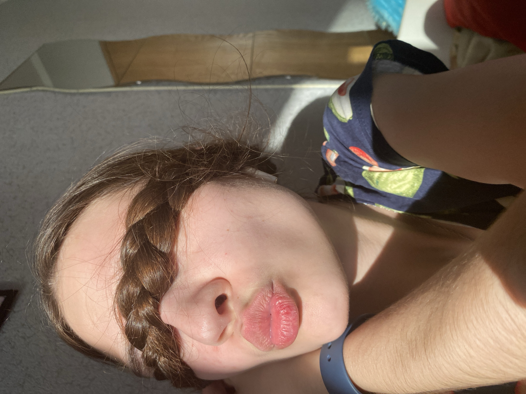 A female lies supine on a light gray surface in a sunlit room. Her face is partially obscured by a brown braid draped across her forehead and nose. She has fair skin and her lips are pursed in a pouting expression. She is wearing a blue smartwatch on her wrist, and a dark blue garment with a green and red pattern is visible near her head. The location is not discernible. There is no evidence of Votkinsk, Russia.