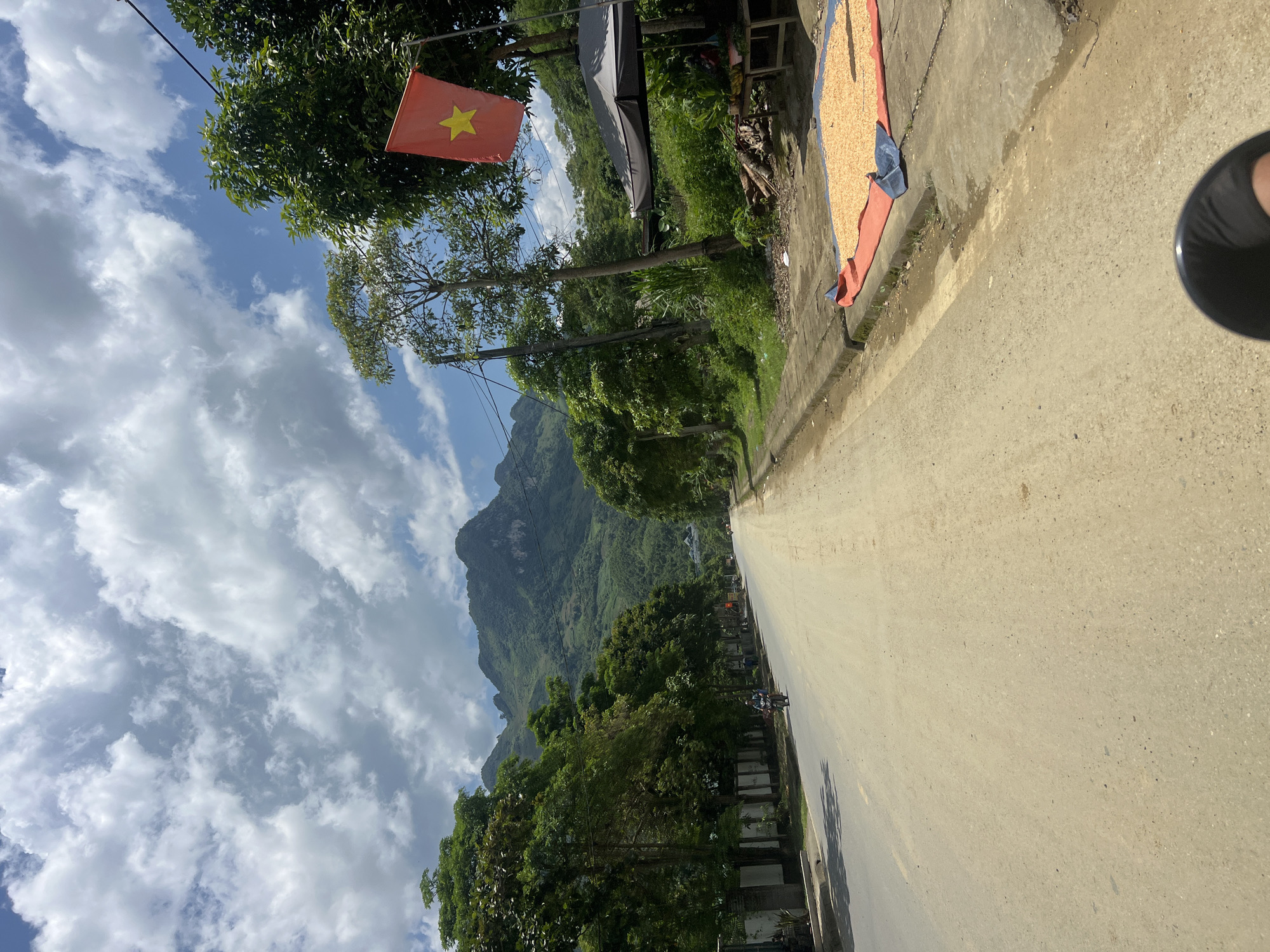 The image depicts a rural scene in Minh Tân Commune, Vietnam, with a paved road leading towards a lush, mountainous landscape. The sky is a vibrant blue, punctuated by fluffy white clouds. On the right side of the road, a red Vietnamese flag with a yellow star flies atop a pole. Next to it, a vendor's stall, covered by a dark umbrella, displays produce on a table. In front of the stall, a large quantity of what appears to be dried corn is spread out on a red and blue tarp on the ground. Several trees line the road and the mountainside, their green foliage contrasting with the dusty road surface. A few buildings are visible on the left side of the road, and a single motorcyclist is riding down the road in the distance. The overall impression is one of a sunny day in a developing rural area, with elements of commerce and daily life visible.