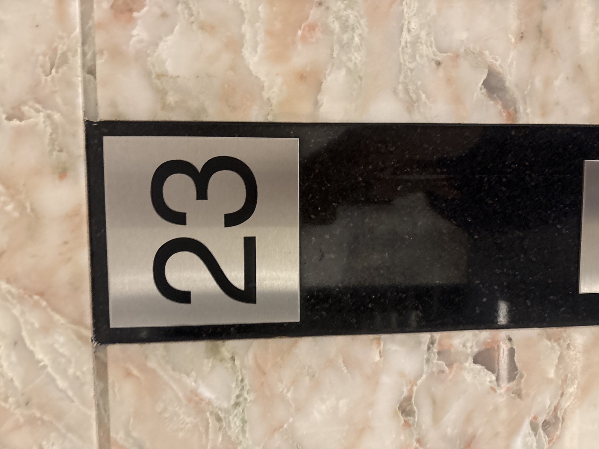 The image shows a close-up of a metallic sign with the number "23" in bold black font. The sign is mounted on a dark, speckled vertical panel, which is itself set against a backdrop of marble with pink and white veining. The marble appears to be a decorative wall finish. There are no people or discernible actions depicted. The setting is a building interior, possibly a hotel or residential complex, given the presence of a floor number. This image is not specific to Dubai, United Arab Emirates.