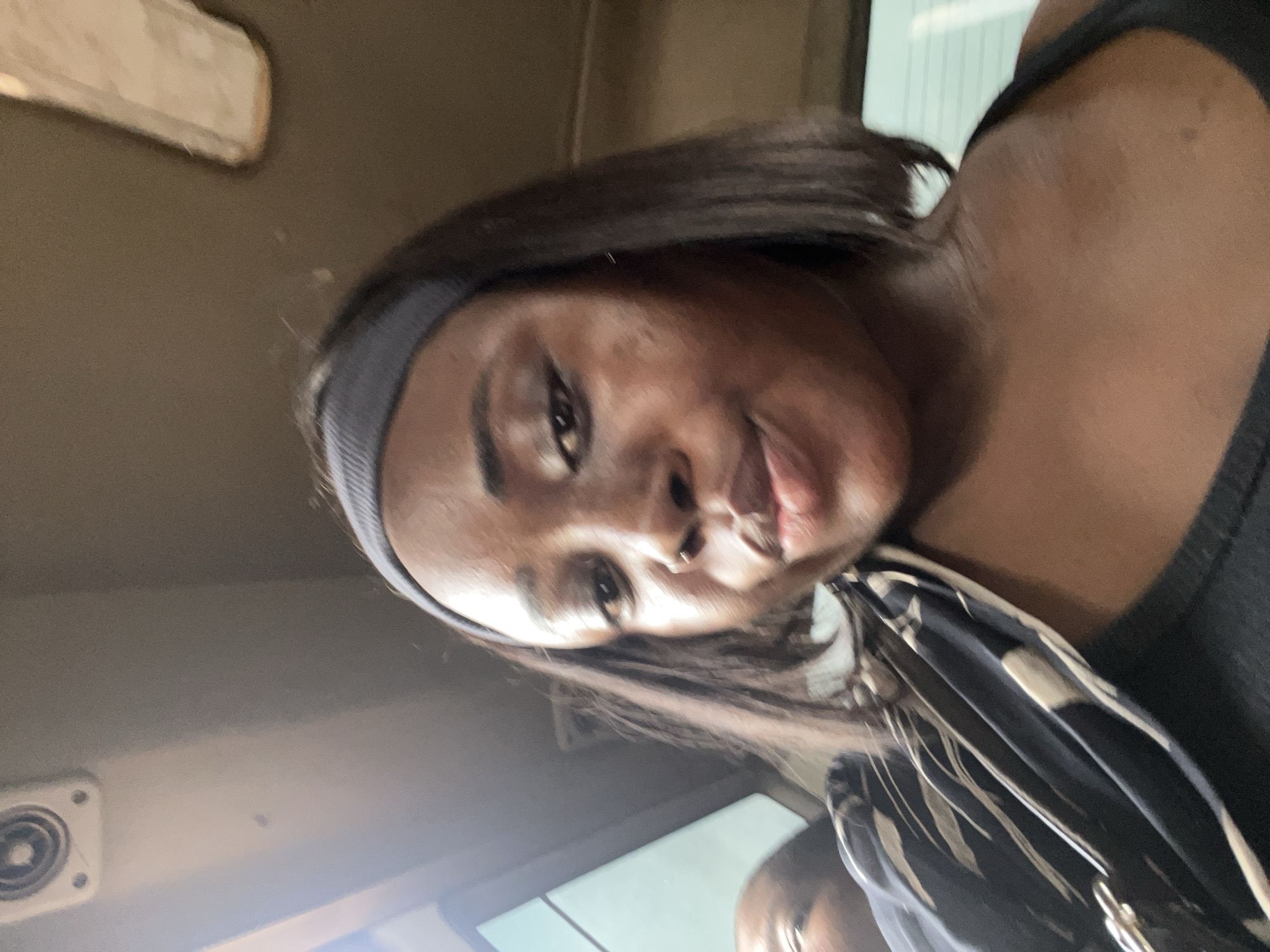 A close-up, vertically oriented selfie shows a Black woman with dark hair, wearing a black tank top and a grey headband. She is looking directly at the camera with a slight smirk. Her skin appears smooth and her eyes are open. She is holding a patterned bag with black and cream stripes. The background is dimly lit and appears to be the interior of a vehicle, possibly a bus, with a beige-colored panel visible in the upper left corner and a circular vent on the left side. The lighting suggests it might be daytime. The image is slightly blurry, contributing to a casual and candid feel. There is no visible text in the image. The subject's expression is neutral to slightly playful.