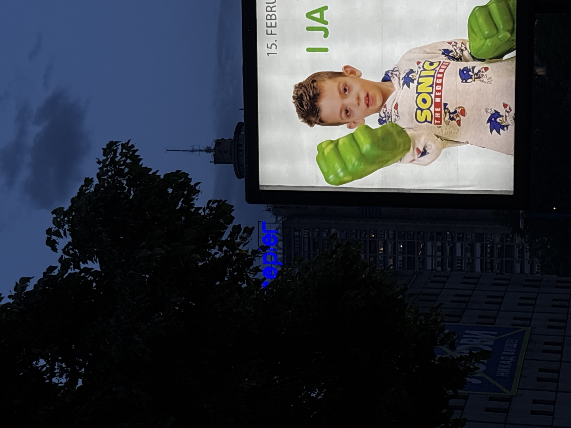 The image depicts an evening scene in Belgrade, Serbia. A billboard displays an advertisement featuring a young boy wearing a Sonic the Hedgehog shirt and green Hulk hands. The text "15. FEBRUAR" and "I JA" are visible on the billboard. In the background, partially obscured by a tree, is the Belgrade TV tower. The building with the blue "Leder" sign, visible behind the billboard, is the building in Terazije number 23, where the "Leder" store is located. Another sign below the billboard reads in Cyrillic, "NIKAD DUŽE!".