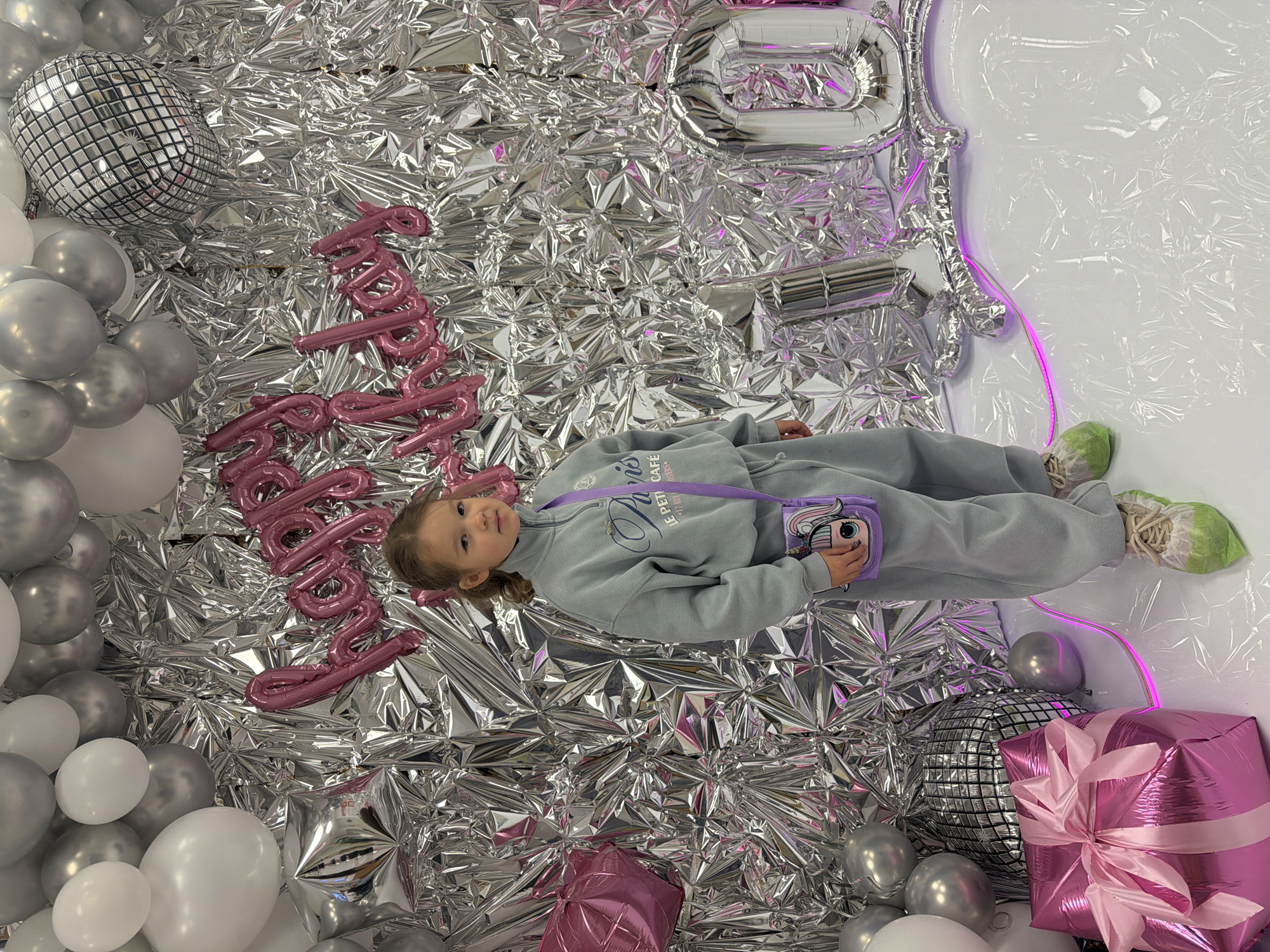 A young Caucasian girl is lying down on a silver, reflective surface, possibly a backdrop for a photograph. She is dressed in a grey sweatshirt and sweatpants, with a small purple purse with cartoon characters on it resting near her hand. Her hair is pulled back and she is looking at the camera.
Around her are decorations that suggest a birthday celebration. There are various balloons, including silver spherical balloons that resemble disco balls, white and silver round balloons, pink rectangular balloons resembling gifts with ribbons, and pink cursive letter balloons spelling "Happy Birthday." There is also a large silver balloon in the shape of the number 10. The backdrop itself appears to be made of reflective material similar to foil, creating a shimmering effect. A neon pink light is visible on the right side of the image, near a section of wrinkled white plastic, which may be part of the studio setup.
There are no explicit indicators of location in Saint Petersburg, Russia. There are no distinctive landmarks, street signs, or architectural details that would point to this specific city.