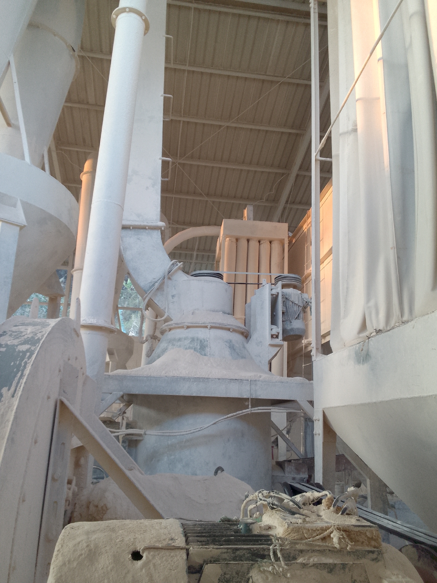 The image shows the interior of an industrial facility in Gunungmasigit, Indonesia, likely a processing plant for minerals or raw materials given the dusty environment and large machinery. Dominating the foreground is a large, conical structure covered in dust, with a significant amount of material piled around its base. To the left, a large, curved metal component, possibly part of a conveyor or processing unit, is also heavily dusted. Several large, white pipes, varying in diameter, are prominent throughout the scene, some vertical and others angled. In the background, a complex arrangement of machinery is visible, including a rectangular unit with multiple cylindrical components and what appears to be an electric motor. The ceiling of the facility, constructed of corrugated metal, is supported by a network of beams. The overall impression is one of an active industrial setting where material processing is ongoing, resulting in a pervasive layer of dust on all surfaces. There are no people visible in the image.