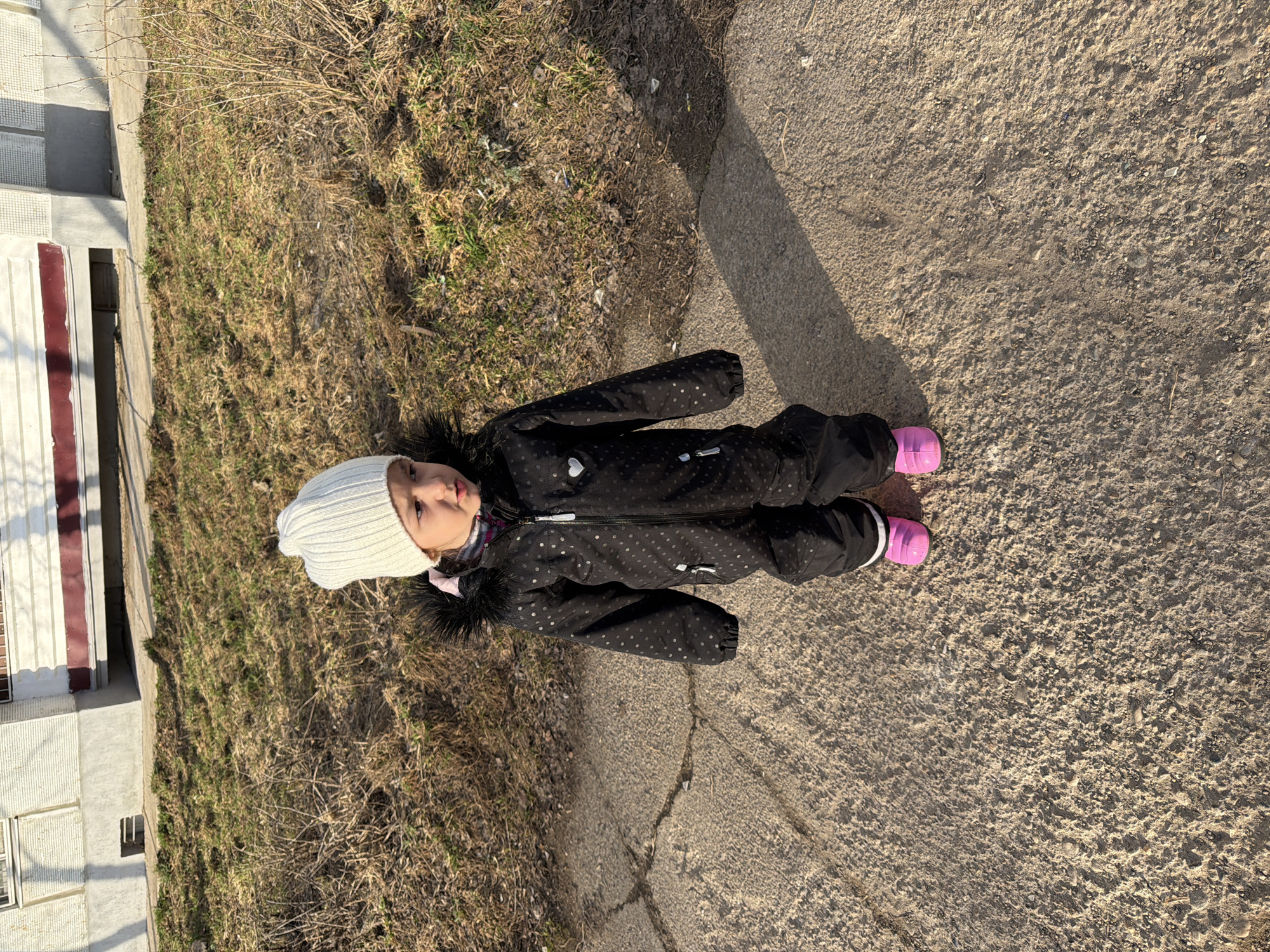 A young girl in a black polka dot snowsuit, white knit hat, and pink boots lies on the edge of a concrete walkway. Her shadow extends from her body onto the walkway. To her left is a grassy area with patches of dead grass. In the background, partially visible, is a light-colored building with a dark red and white accent.
Based on the available information, it is not possible to confirm if this image was taken in the city of Chaikovsky, Russia, or to provide details about the environment in this area.