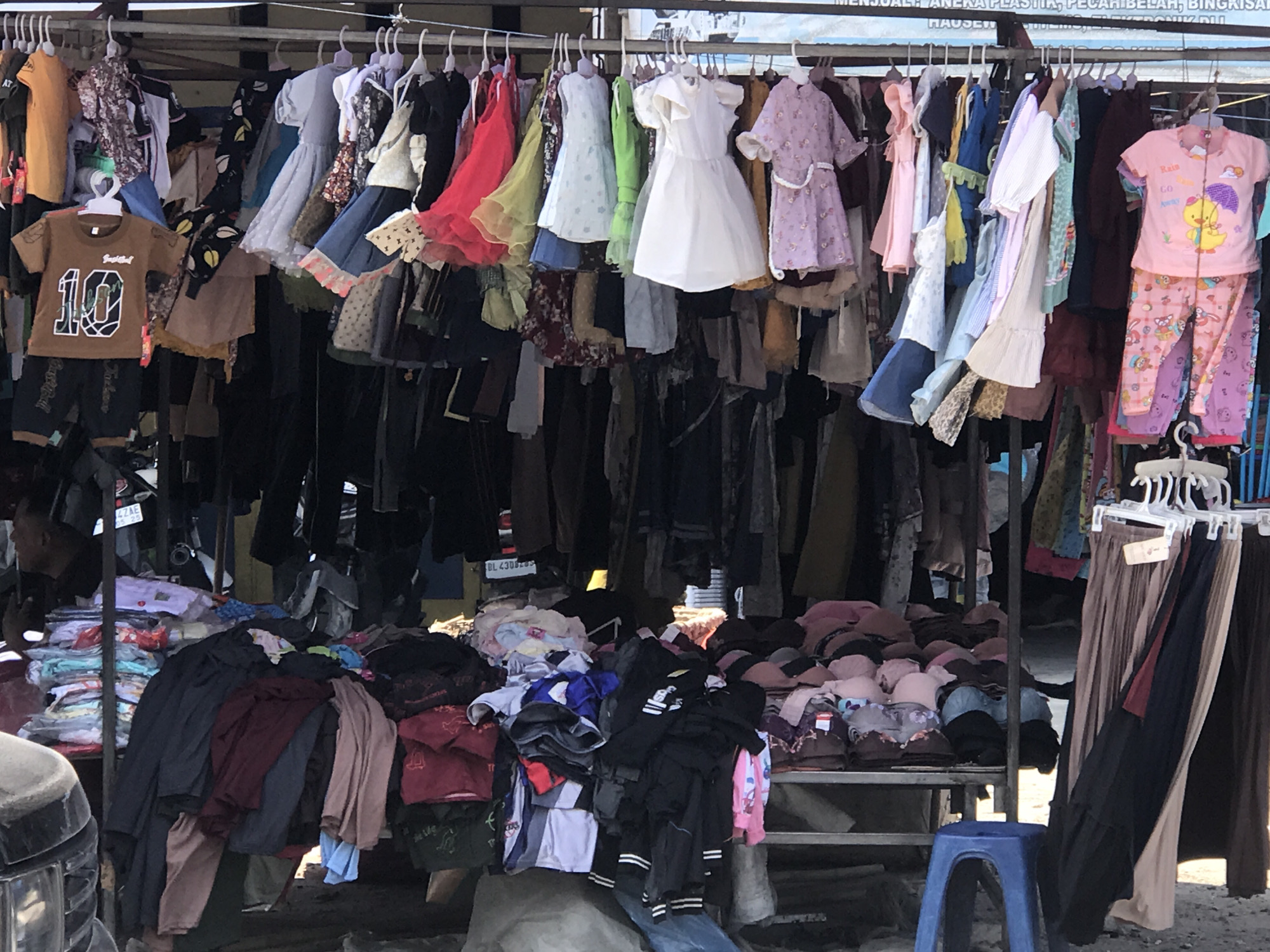 The image captures a roadside clothing stall in Tingkeummanyang, Indonesia. Garments hang densely from metal rods supported by metal posts. The stall has a sign that reads "MENJUAL ANEKA PLASTIK, PECABEHIRUNIK BIU HAUSE". The clothing includes children's dresses, shirts, and pants, hung on plastic hangers. Below the hanging clothes are piles of folded and stacked clothing, including pants, shirts, and what appears to be undergarments. A small blue plastic stool sits on the ground near the right edge of the frame. A vehicle is partially visible on the left side of the frame. The area surrounding the stall appears to be a paved roadside.