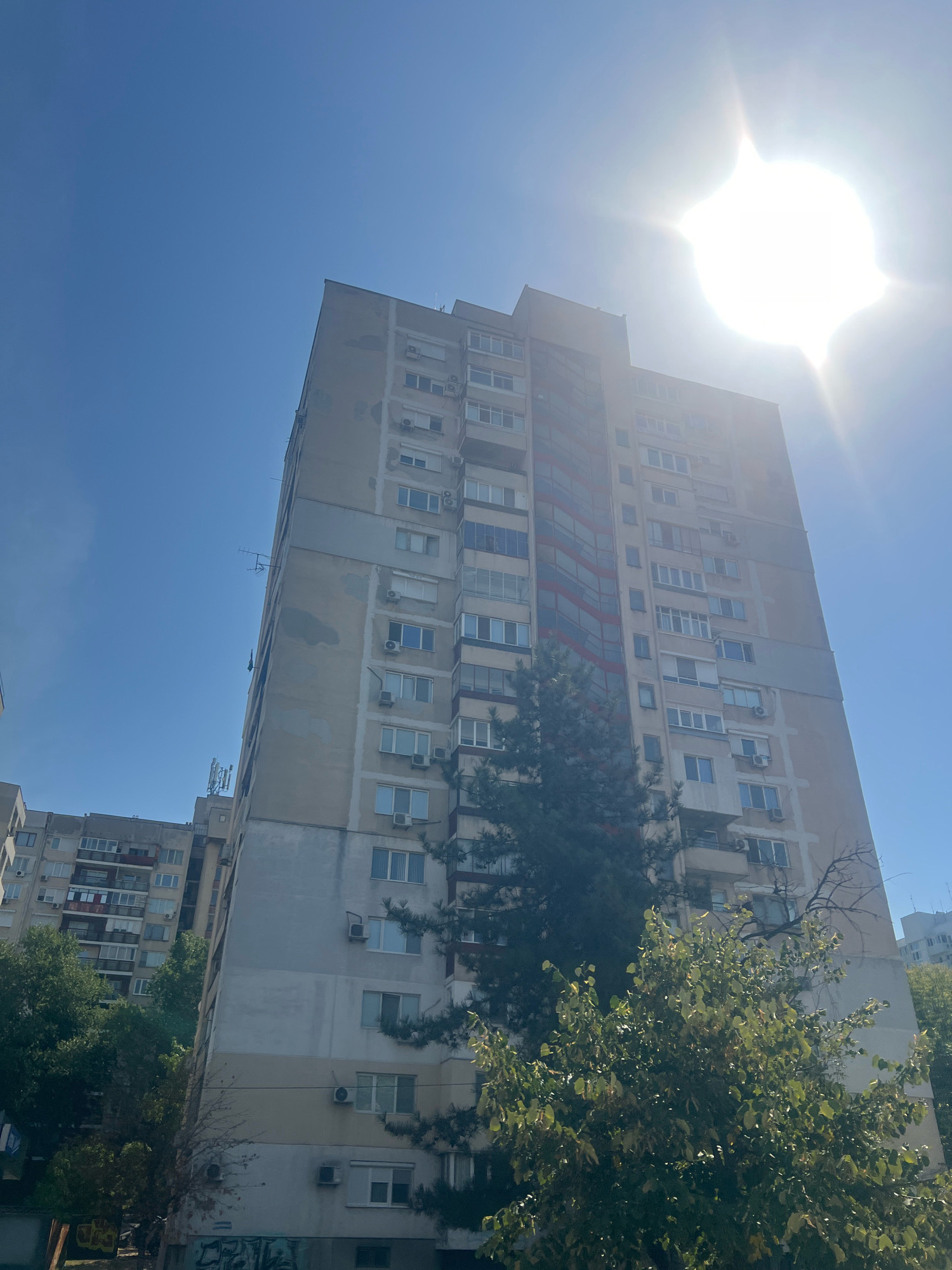 The image shows a tall, multi-story apartment building under a bright, sunny sky with the sun flaring prominently in the upper right quadrant. The building, appearing somewhat aged with visible wear on its facade, is made of concrete and has numerous windows and air conditioning units. There are trees in the foreground, partially obscuring the lower portion of the building. To the left, another apartment building is partially visible. The scene appears to be in a residential area of Plovdiv, Bulgaria. There are no people or discernible actions depicted.