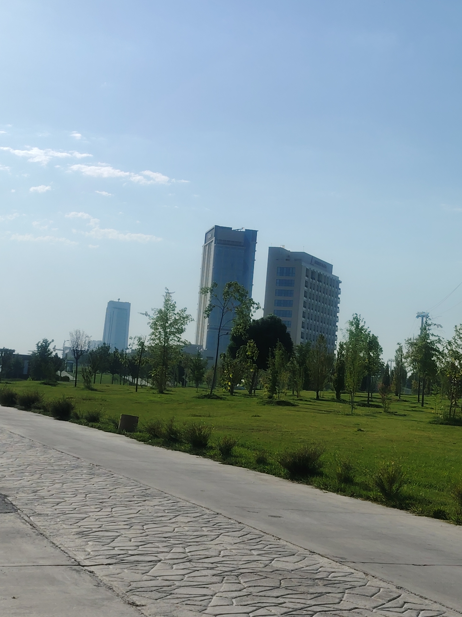 The image shows a paved walkway in the foreground, leading into a grassy park area. Several trees are scattered throughout the park, and in the background, there are modern high-rise buildings. One building appears to be a hotel, with "SKYPLAZA" visible on its facade. The sky is blue with some scattered clouds. The scene appears to be a public park or urban green space in the city of Konigil, Uzbekistan, likely taken during daylight hours. There are no people or animals visible in the image. The overall impression is one of a modern urban environment with well-maintained green spaces.