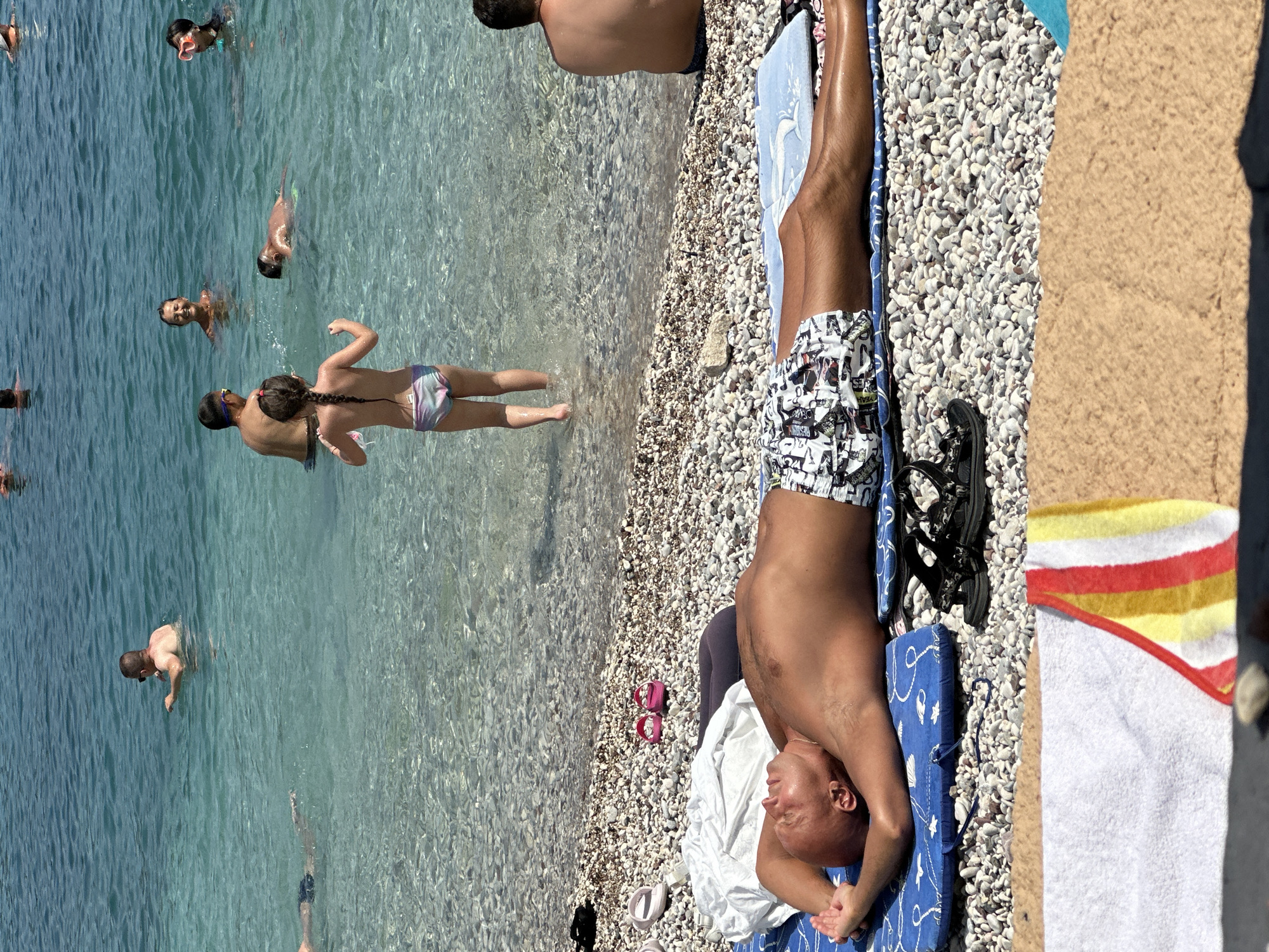 The image depicts a sunny day at a pebble beach in Budva, Montenegro. In the foreground, a bald man is lying on his back on a blue mat, with his hands behind his head. He is wearing patterned swimming shorts and appears to be sunbathing. Adjacent to him are black sandals and a white towel folded next to a dark item of clothing. In the middle ground, several people are visible in the clear turquoise water. A young girl with braided hair stands in the shallow water facing away from the camera. Further out, a woman smiles in the water, and a child with goggles is swimming. In the background, other individuals are enjoying the sea, with the clear water revealing the rocky seabed. The overall scene conveys a relaxed beach atmosphere.