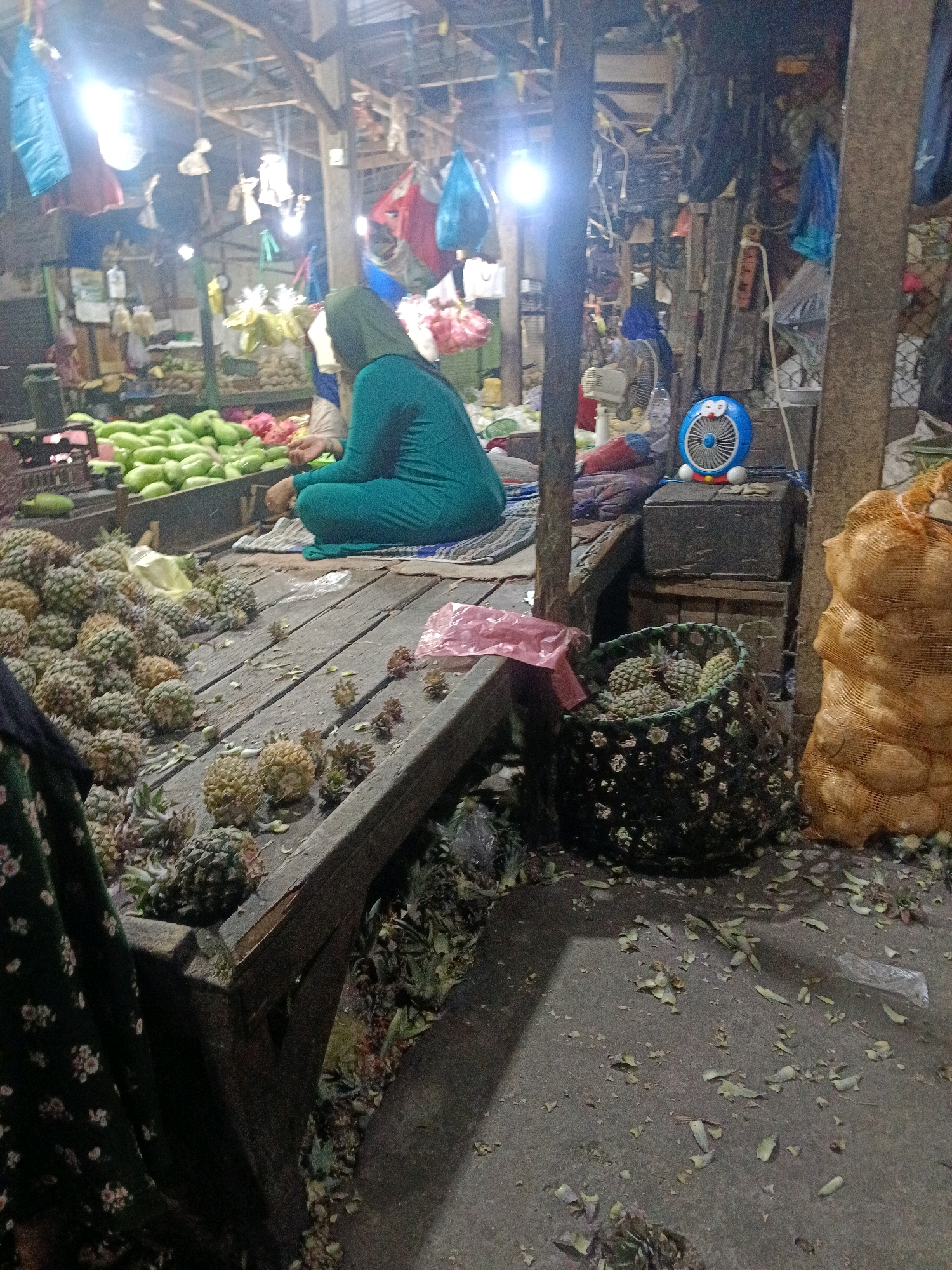 In the city of Wonocolo, Indonesia, a market scene unfolds. A woman, dressed in a teal hijab and matching long-sleeved garment, is seated on the wooden floor, facing away from the camera. She appears to be a vendor, with a pile of green gourds or gourds in the background. In the foreground, a large collection of pineapples is displayed on a rustic wooden surface. Some pineapples are loose, while others are contained within a woven basket. The market environment is dimly lit, with overhead lighting casting shadows. Various other produce and goods are visible in the background, along with hanging bags and what appears to be a blue Doraemon-themed fan. The ground is covered with scattered leaves and debris, indicative of an active marketplace.