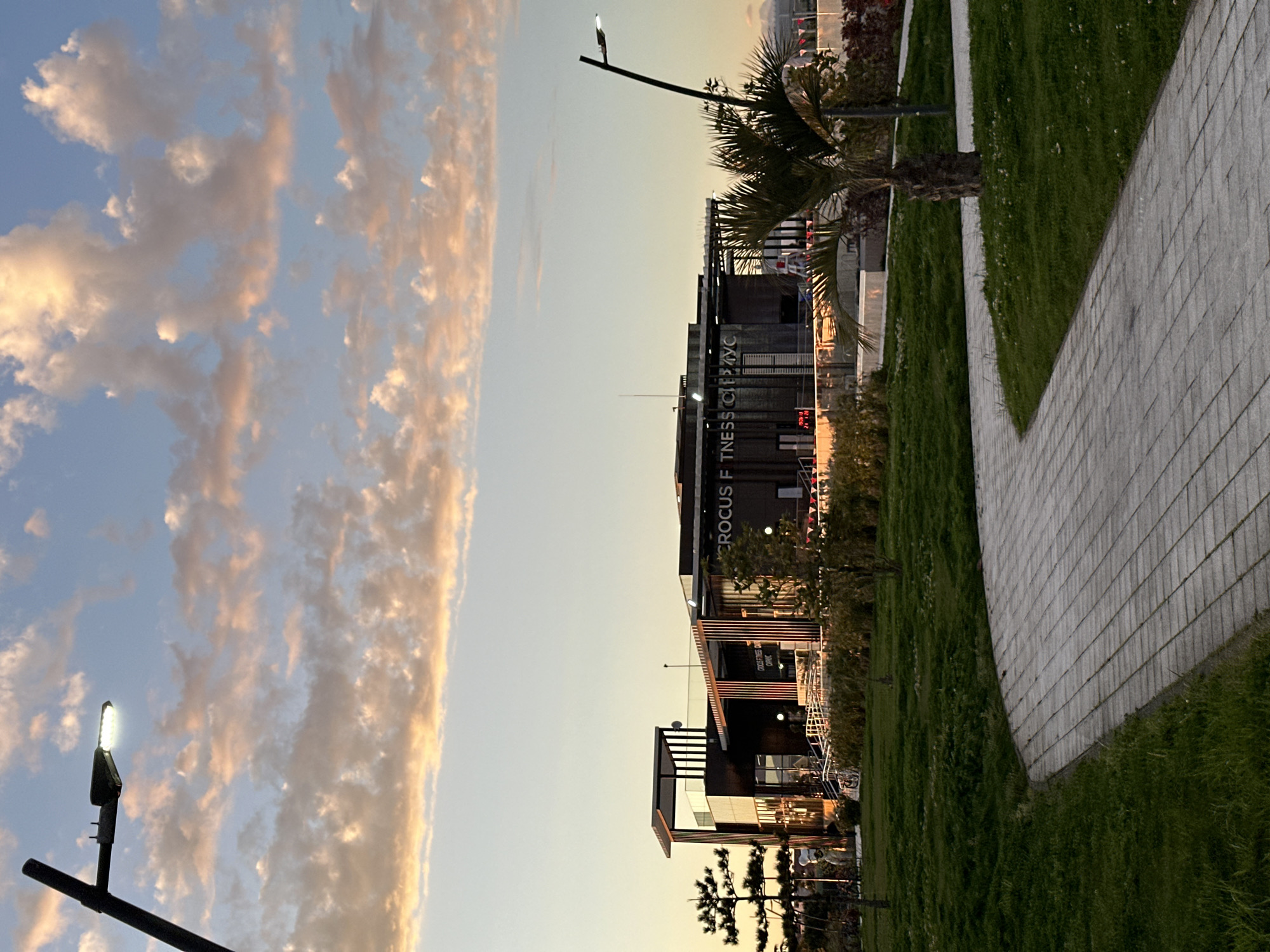 The image shows an outdoor scene in the city of Sirius, Russia. The sky is visible with a mix of blue and light orange clouds. A tall building with the words "CROCUS FITNESS CITY" in English is present. To the right of the building is a palm tree and grass. A paved walkway runs alongside the grass. A lamppost is visible on the left side of the image and another is visible near the building.