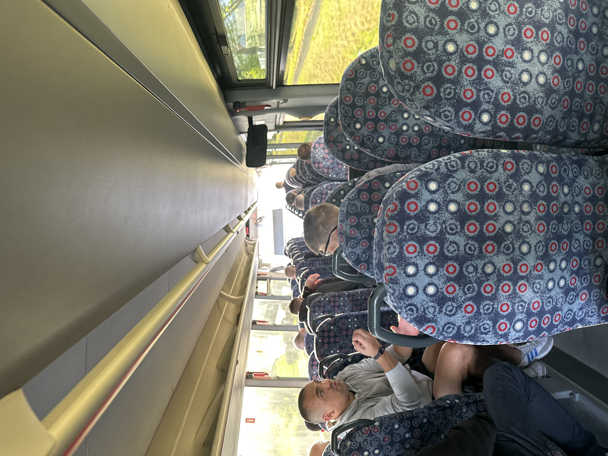 The image depicts the interior of a bus, likely en route to or from Marcinowice, Poland, based on its context. The perspective is from a passenger seated on the right side, looking forward and slightly to the left. The aisle is visible, lined with seats upholstered in a patterned fabric of blue, grey, red, and white circles. Several passengers are seated, with most facing forward.
In the foreground, on the left, a man with a shaved head and a grey hoodie is speaking, gesturing with his hands. He is seated next to a person whose legs and lower torso are visible, wearing dark trousers and athletic shoes. To his right, in the row ahead and slightly further back, another man with glasses is visible from the back of his head. Further down the aisle, other passengers are seated in varying degrees of visibility.
The bus interior features overhead compartments and lighting. Large windows run along the right side, offering a view of a green, grassy area with trees and sunlight filtering through. The overall lighting within the bus is natural, coming from the windows, and appears to be daytime. The atmosphere seems casual, with passengers engaged in conversation or simply traveling.