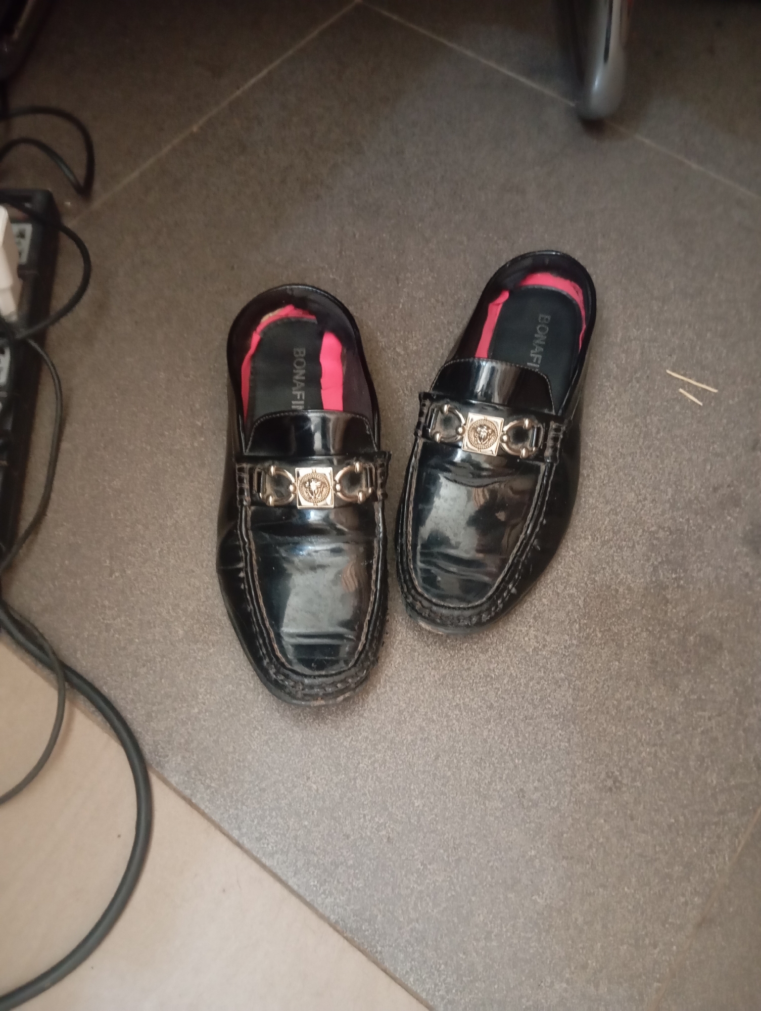 Two black, glossy slip-on loafers with gold accents are positioned on a tiled floor. The left shoe is slightly angled towards the viewer, while the right shoe is more directly facing forward. Both shoes feature a prominent gold emblem on the vamp, which appears to be a stylized Medusa head within a rectangular frame, reminiscent of Versace designs. The inner lining of the shoes is a vibrant pink, and the brand name "BONAF" is visible on the insole of the right shoe. The floor is a textured, dark grey tile, with lighter grey grout lines. To the left of the shoes, a black electrical cord is visible, coiled on a light-colored surface. In the background, a portion of a metallic object, possibly furniture, is out of focus. The scene is lit from above, casting soft shadows. The image appears to be taken indoors, in what could be a residential or commercial setting. The overall impression is one of a static display of footwear. The presence of electrical cords suggests an urban environment, and given the prompt's request to include the location of Jos, Nigeria, this scene could be interpreted as an interior shot within that city.